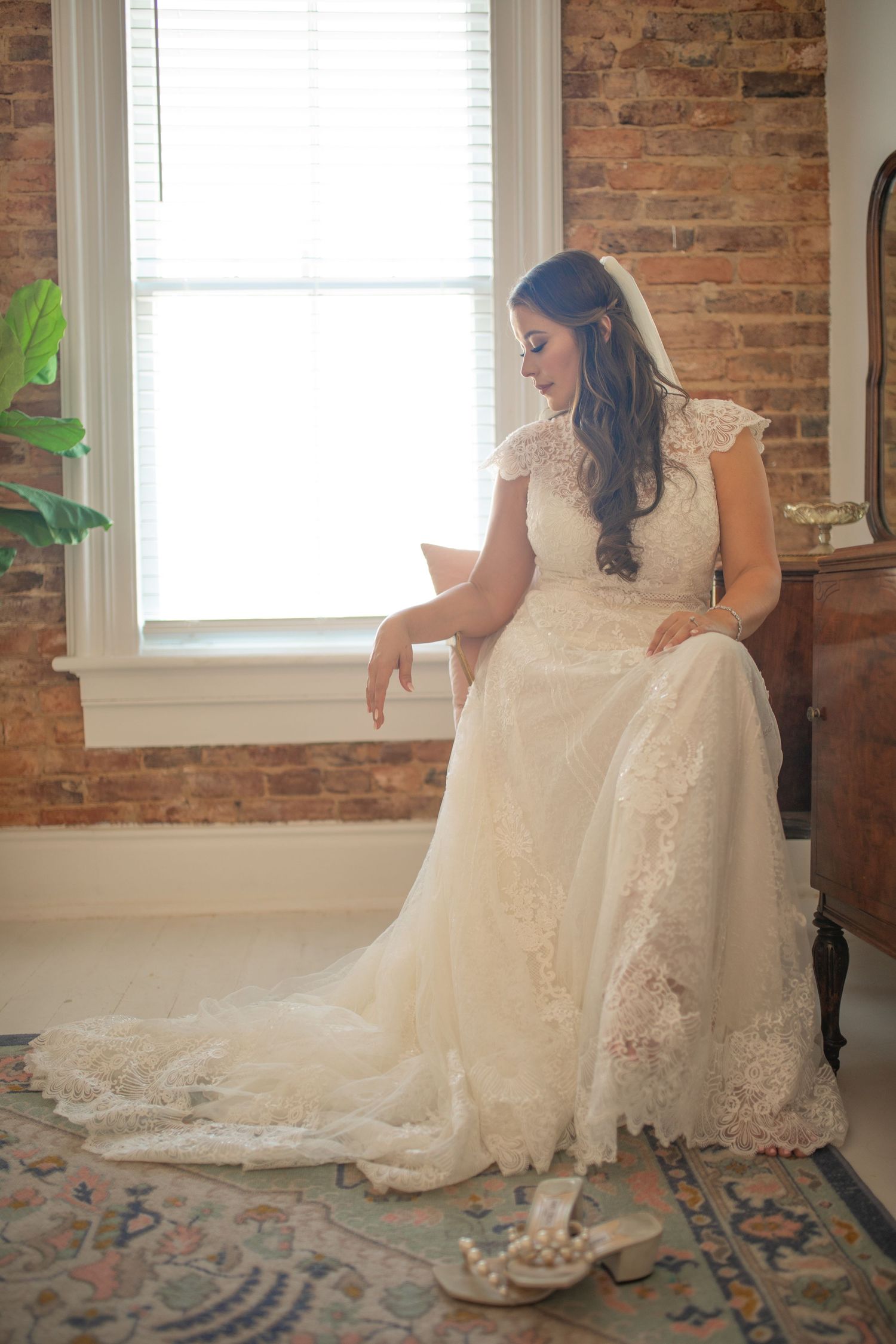 A bride in a lace white wedding dress and sneakers poses near a window with exposed brick wall and vintage carpet.