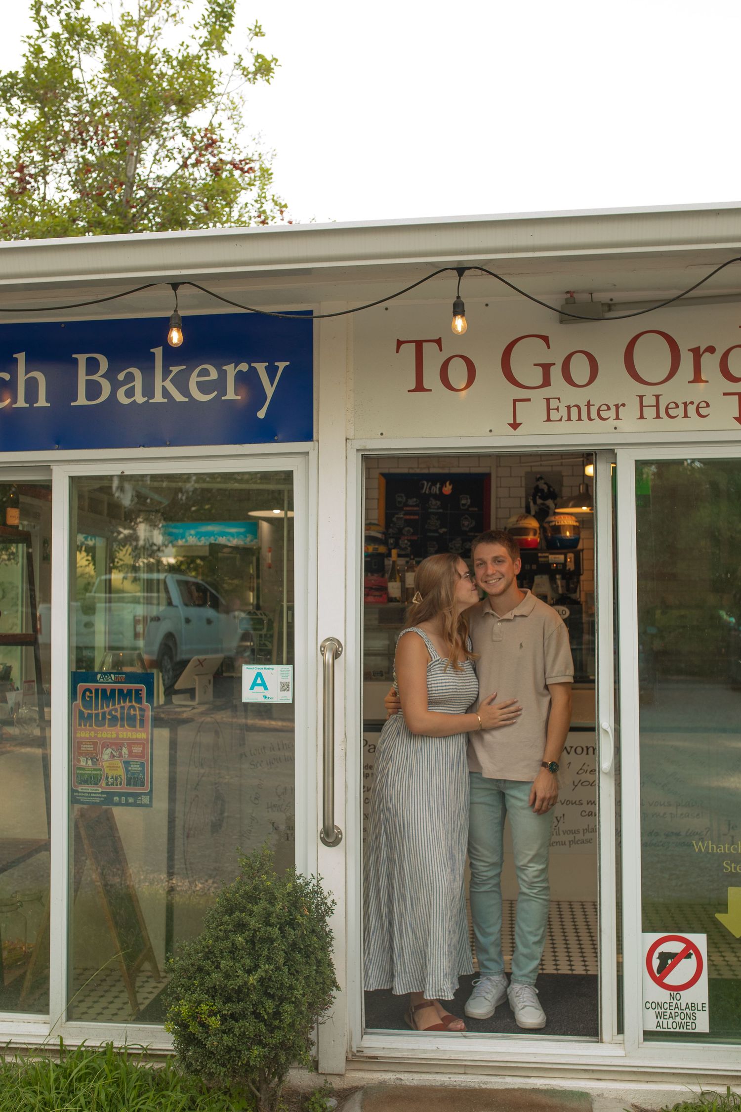 A couple stands in the doorway of a bakery with 'To Go Orders' signage on the exterior window.