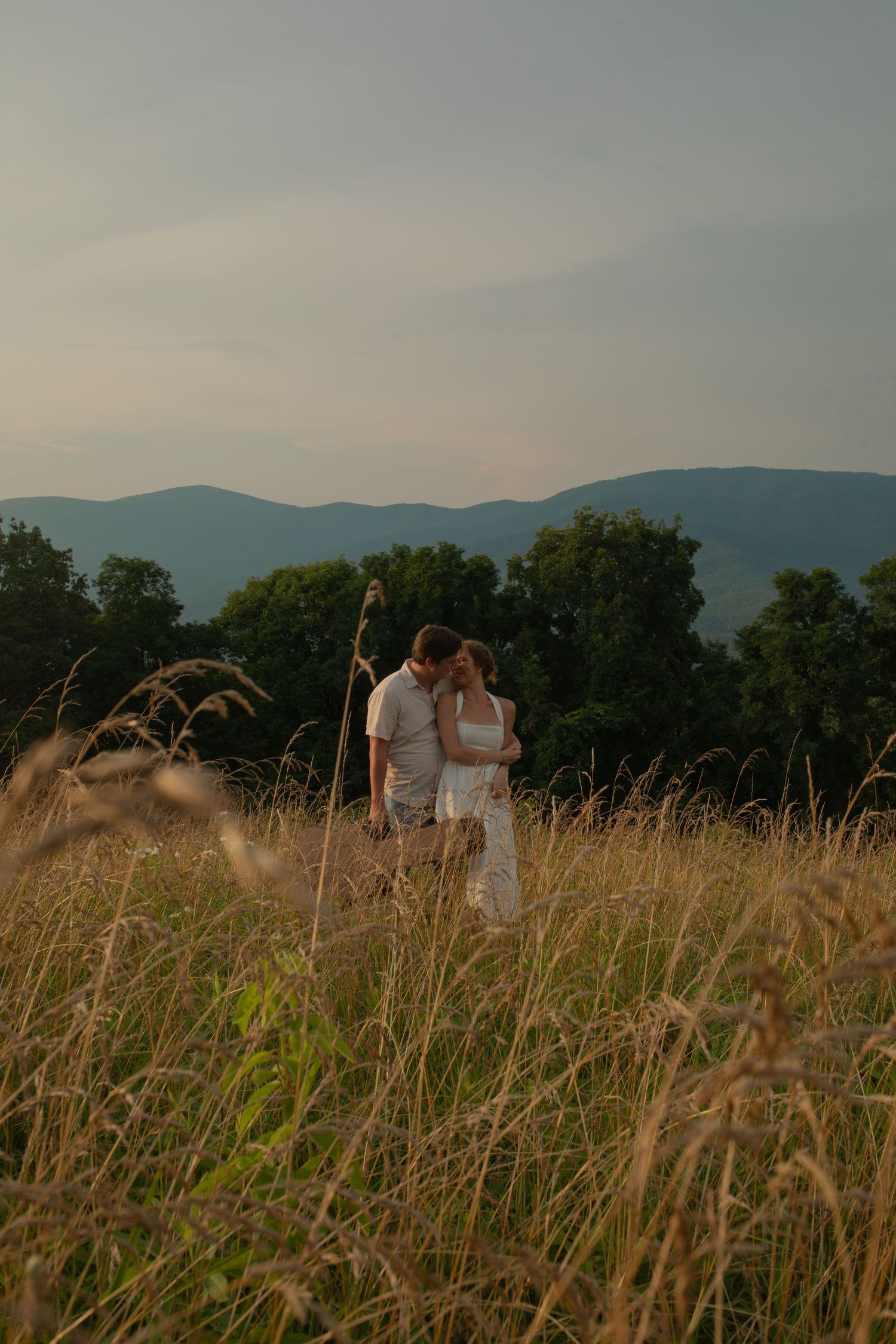 Couple embracing in golden meadow with mountain backdrop at sunset.