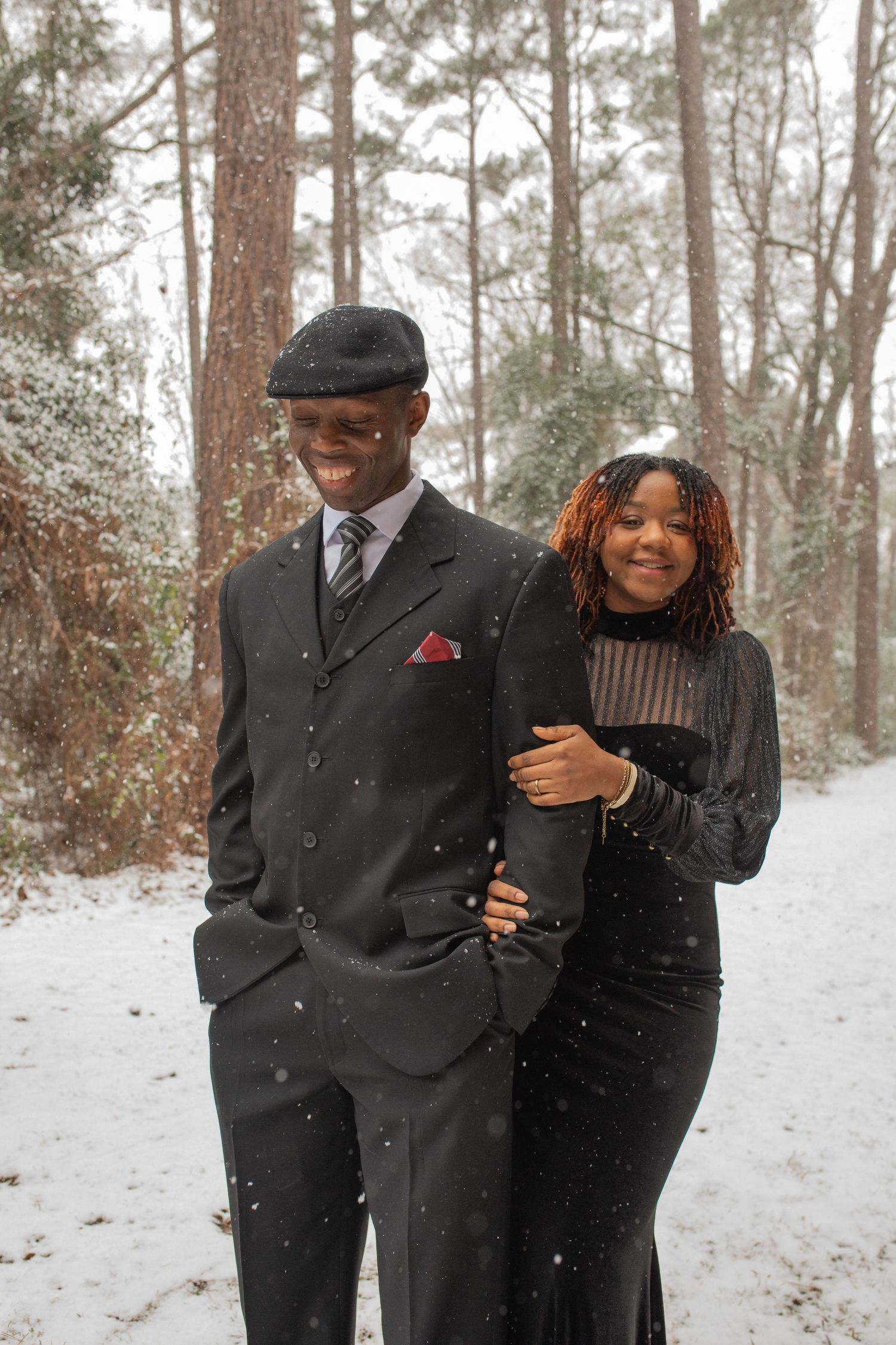 Smiling couple in formal black attire posing together in a snowy forest setting during light snowfall.