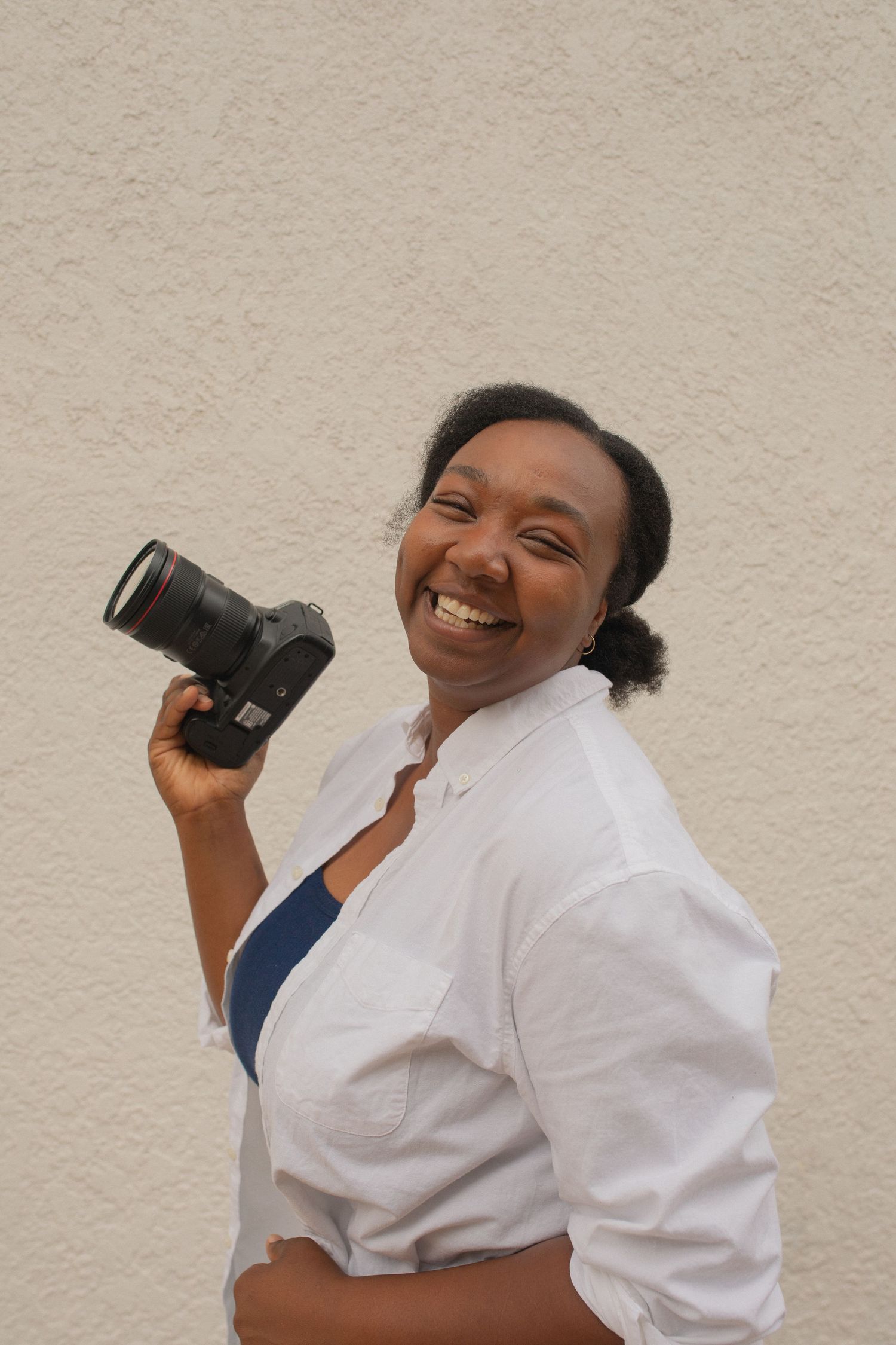 Smiling woman holding a DSLR camera, wearing a white shirt, standing against a light textured wall.
