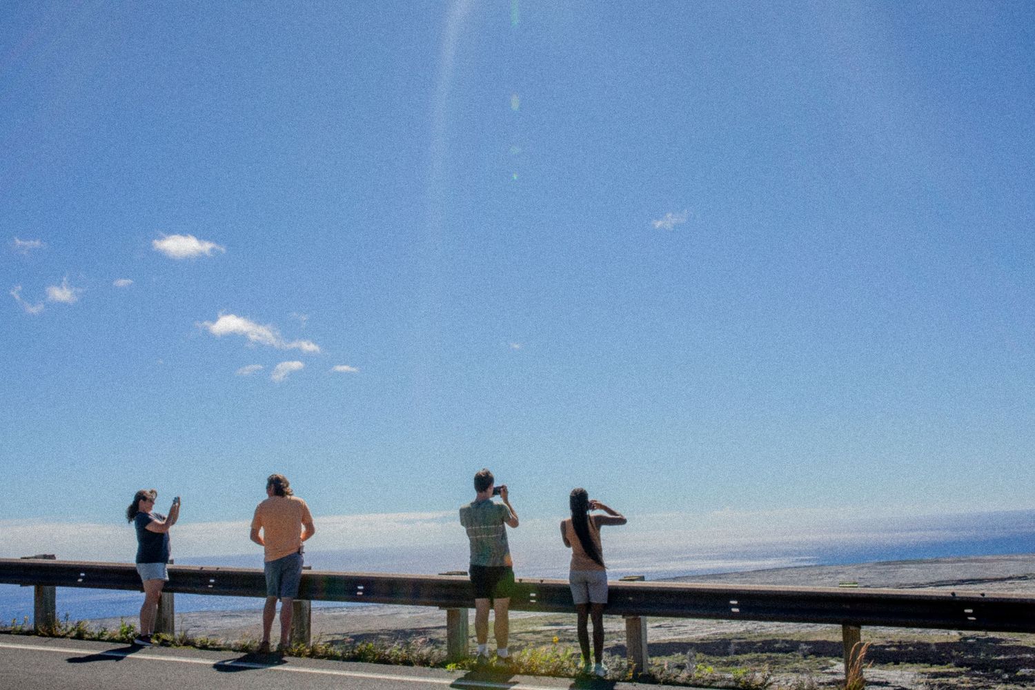 Silhouetted figures stand on an observation deck overlooking a scenic mountain landscape on a clear blue day.