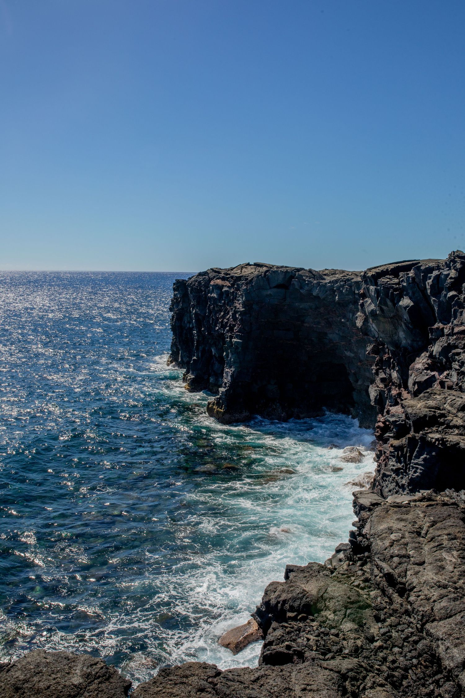 Dramatic volcanic cliffs rise above the crashing blue waves of the ocean against a clear sky background.