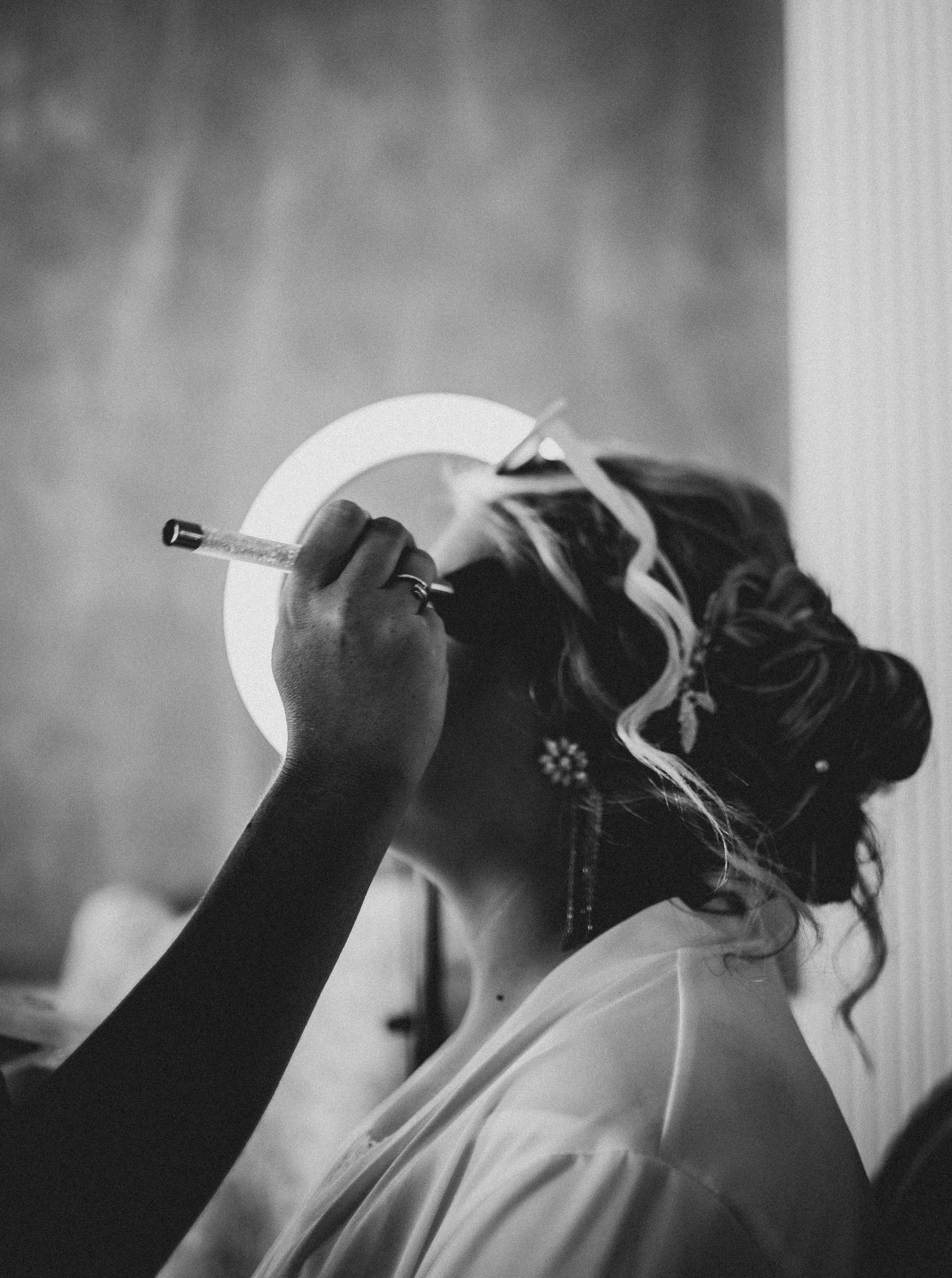 Black and white photo of someone in profile wearing white getting their makeup applied while smoking a cigarette.