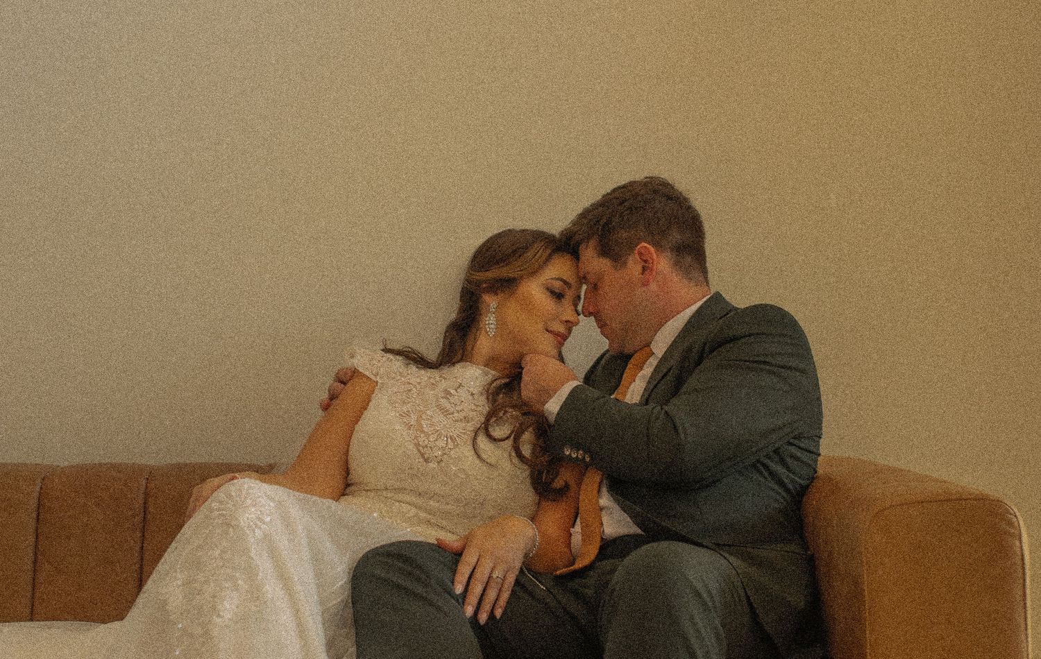 A romantic moment between newlyweds sitting close together on a brown couch in warm lighting.
