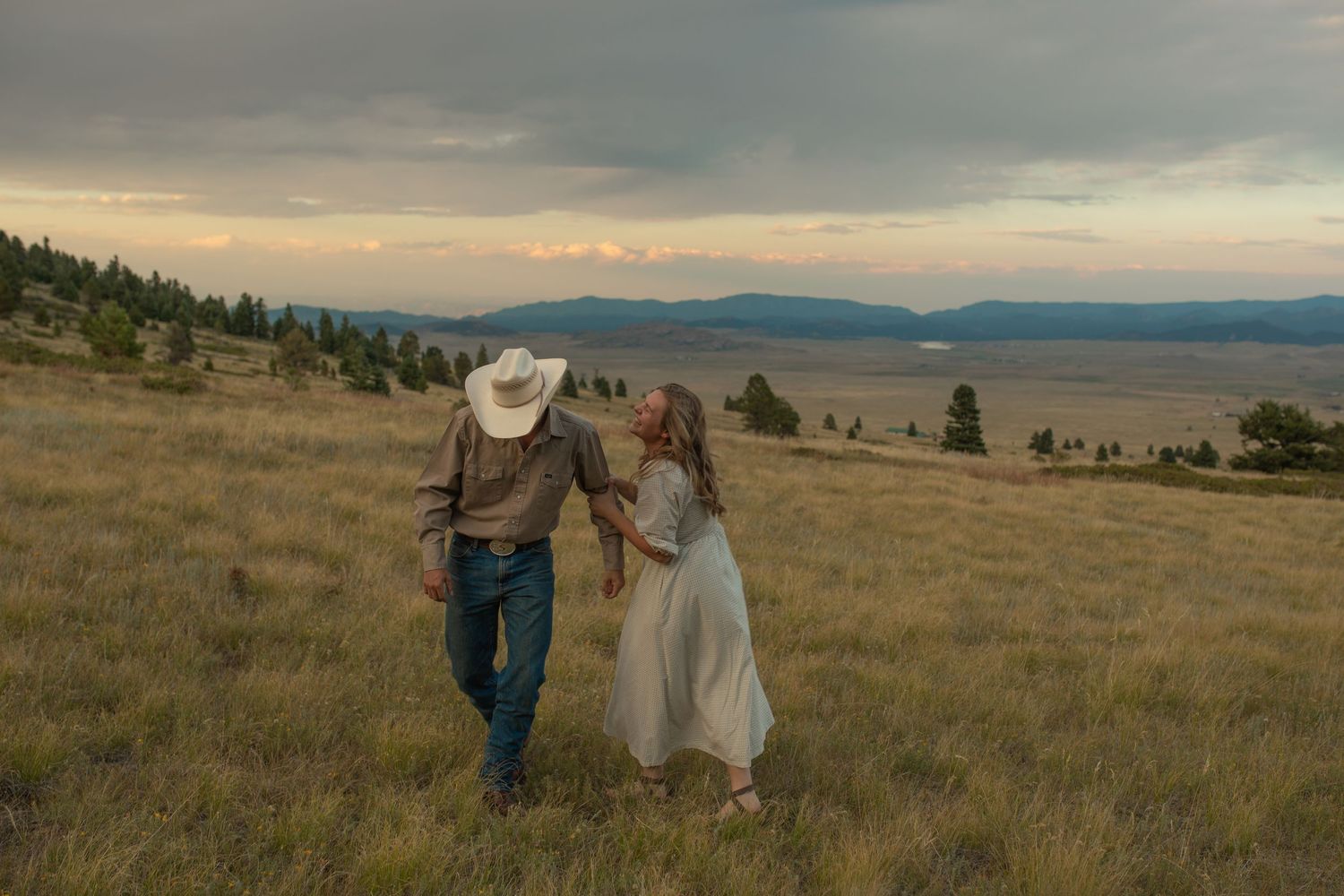 Two people walk hand in hand through a golden prairie field at sunset with mountains in the distance.