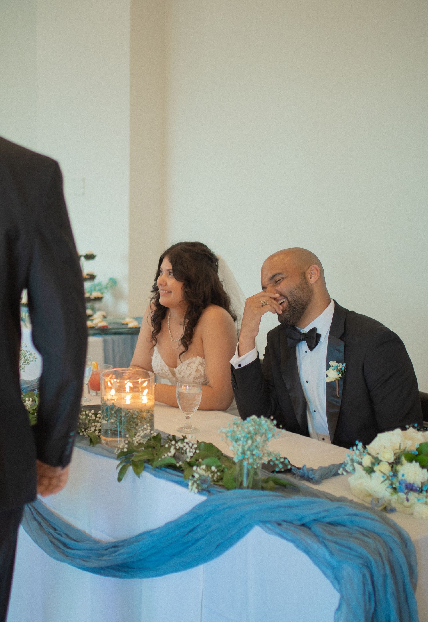 Bride and groom laughing at sweetheart table with blue draping and floral centerpieces during wedding reception.