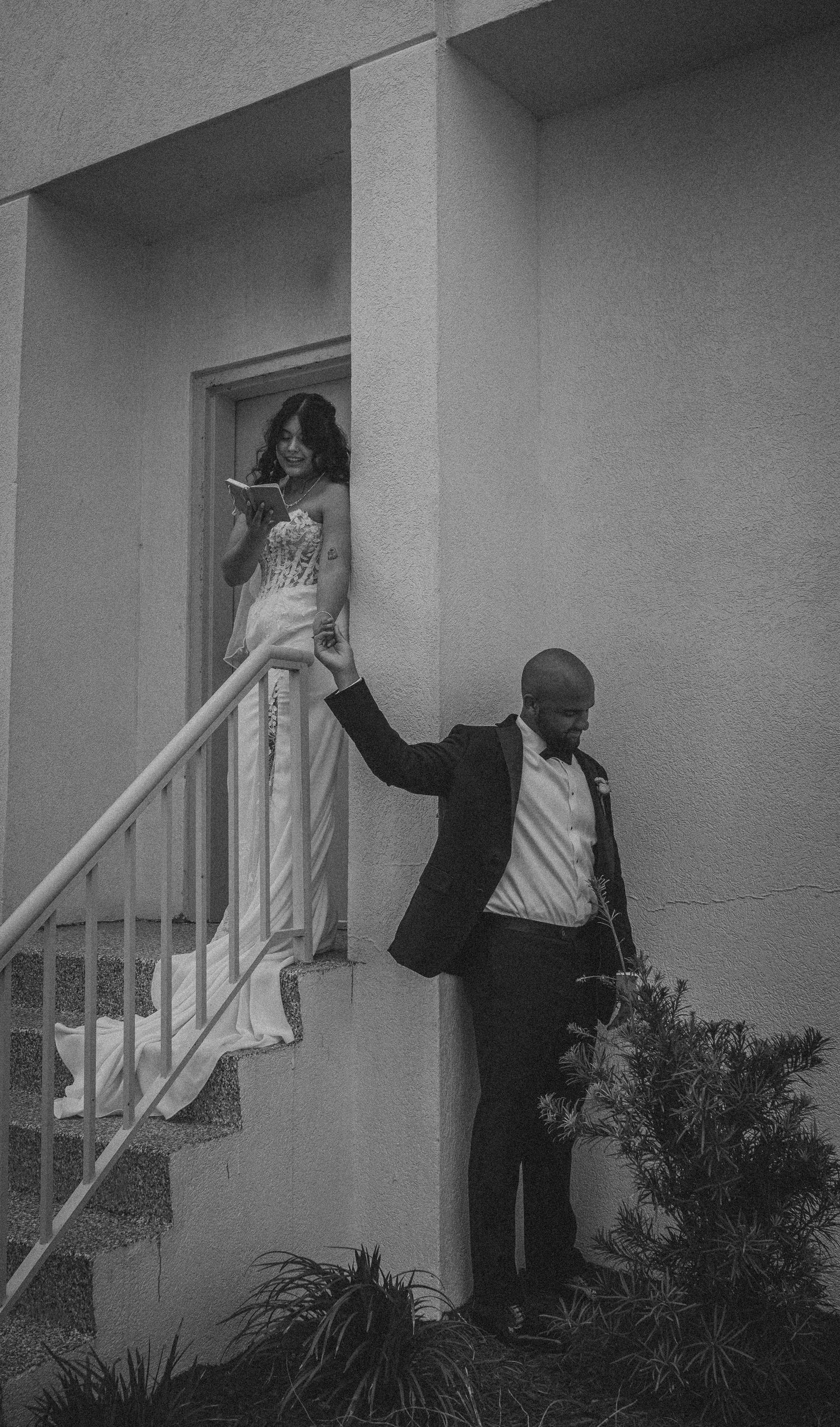 Black and white photo of a bride in a white gown and groom in a tuxedo on outdoor stairs at their wedding.