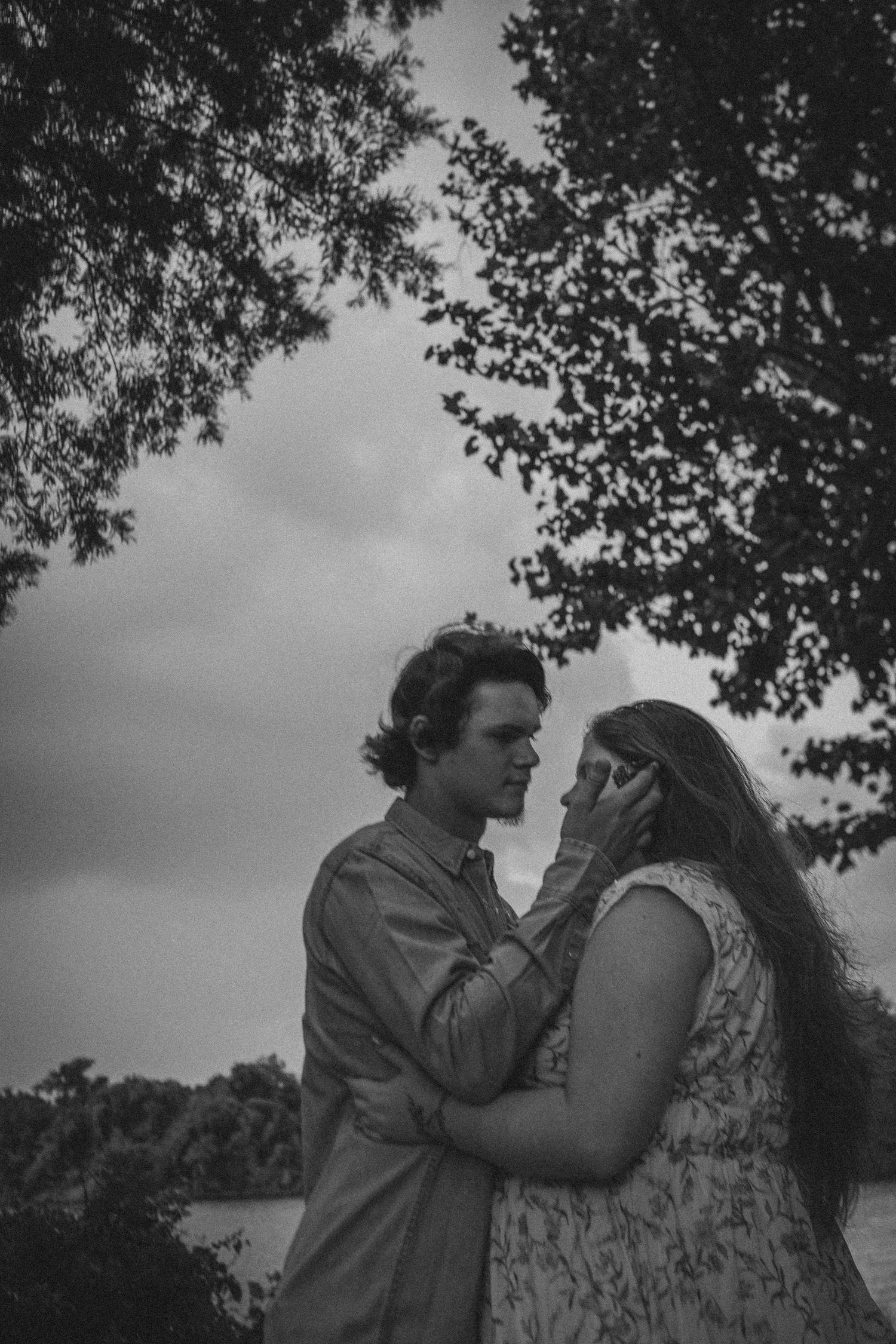 Black and white photo of a romantic couple embracing tenderly under trees with cloudy sky backdrop.