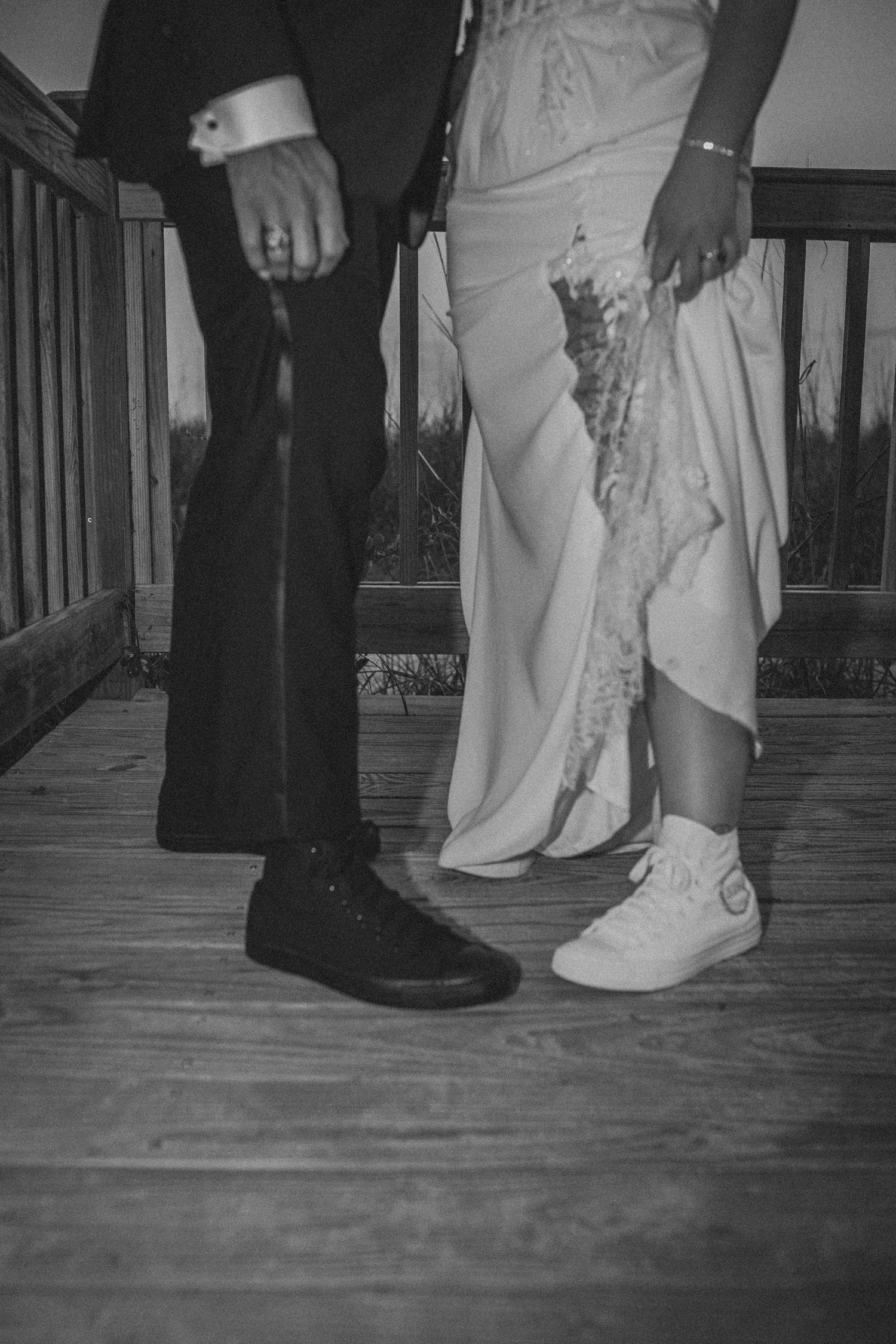 Black and white photo of wedding couple showing groom in black Converse and bride in white Converse sneakers on wooden deck.