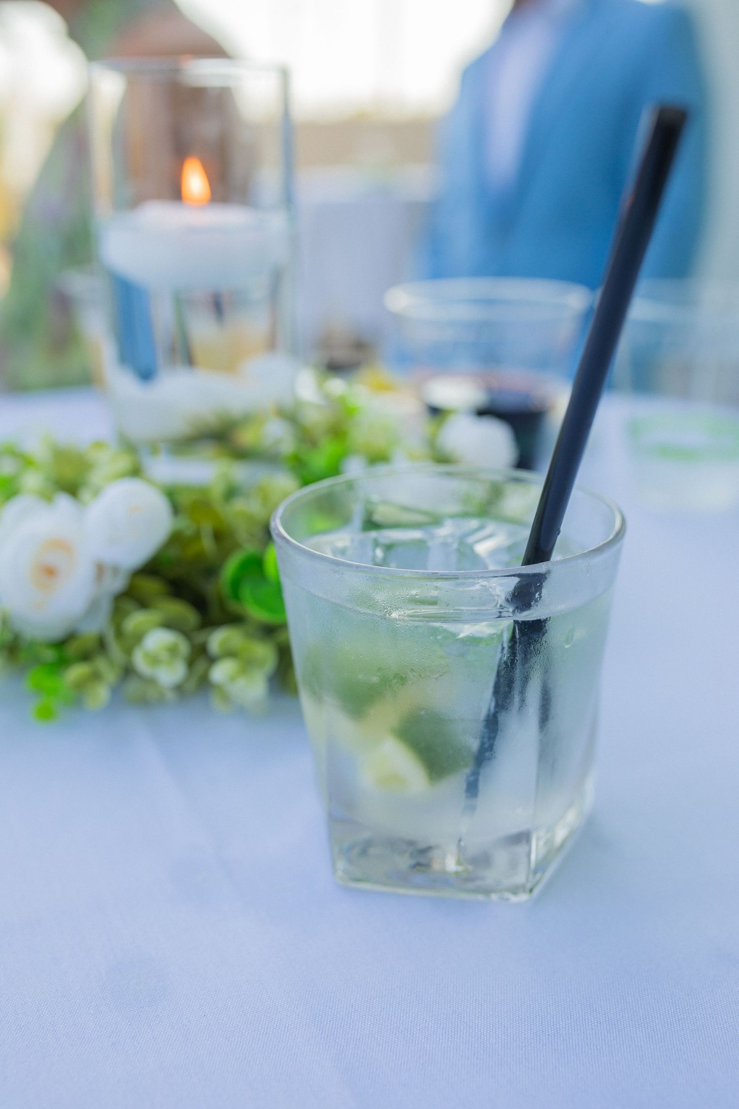 A refreshing mojito cocktail with ice, lime, and a black straw on a white table at an outdoor wedding reception.