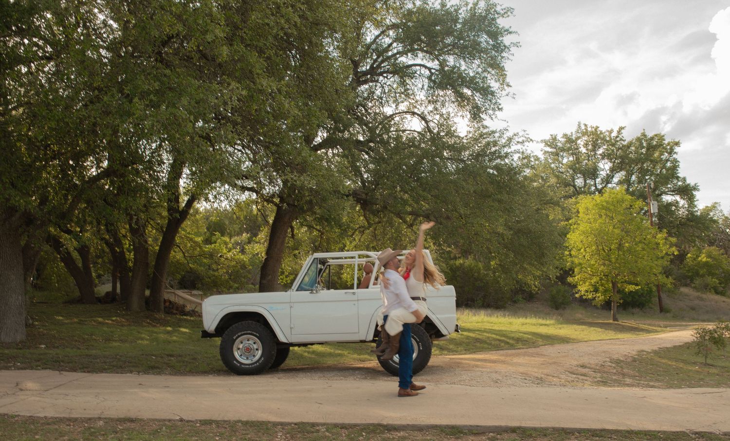 Couple embracing beside a vintage white Ford Bronco on a tree-lined gravel path during a romantic outdoor photoshoot.