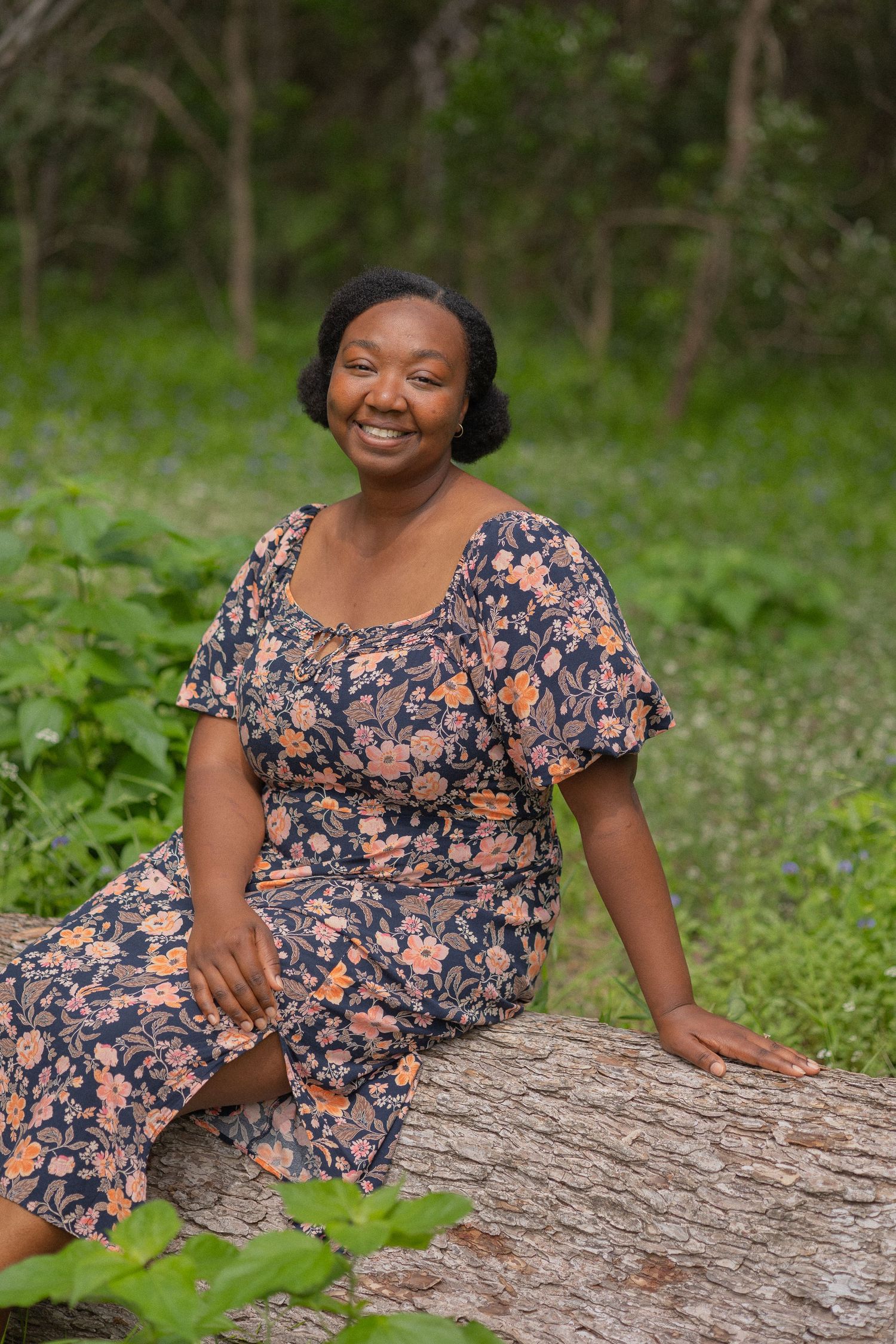 Smiling woman in floral dress sitting on a log in a lush green forest setting.