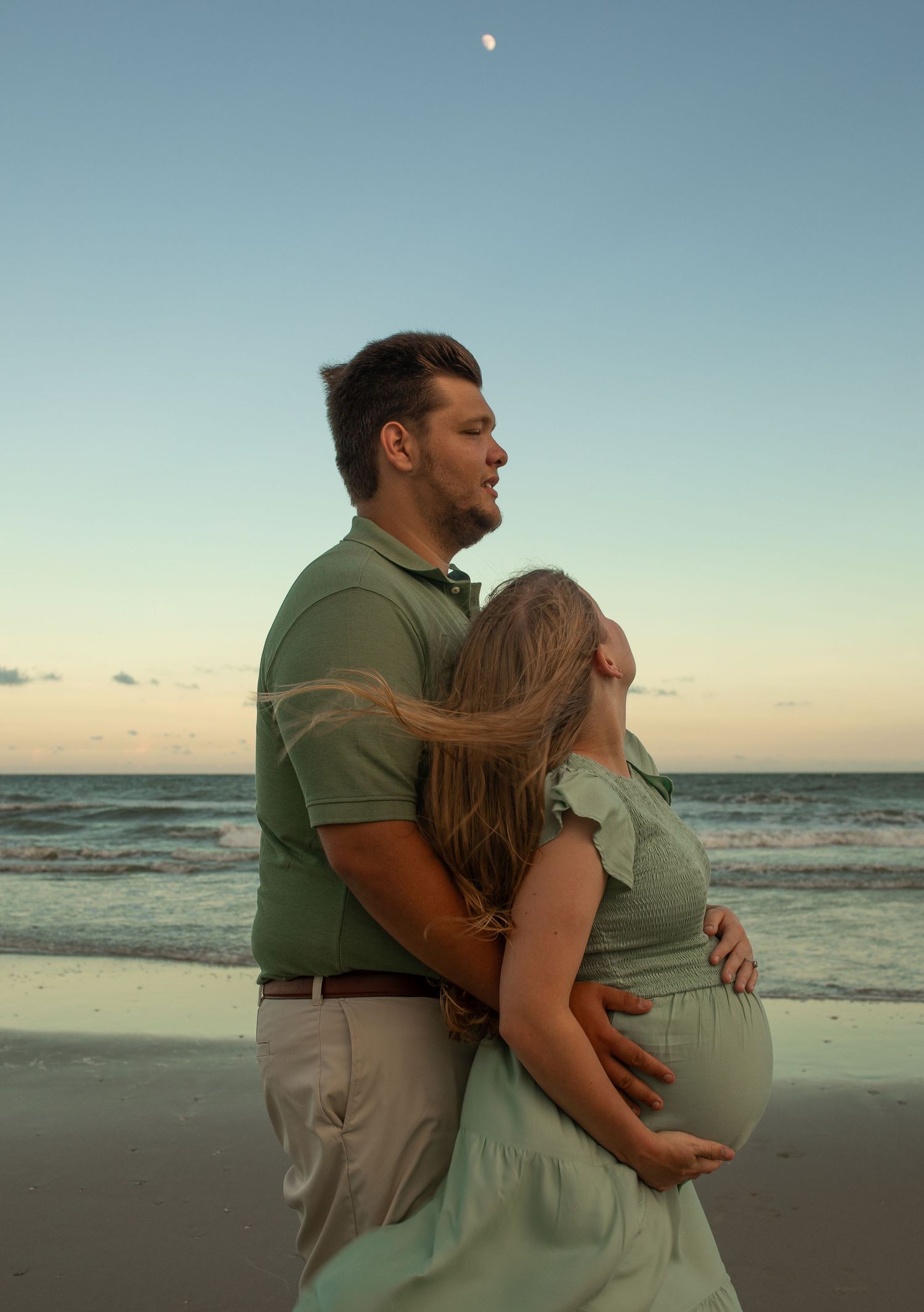 A romantic maternity photo shows a couple embracing on the beach at sunset with the moon visible in the sky.