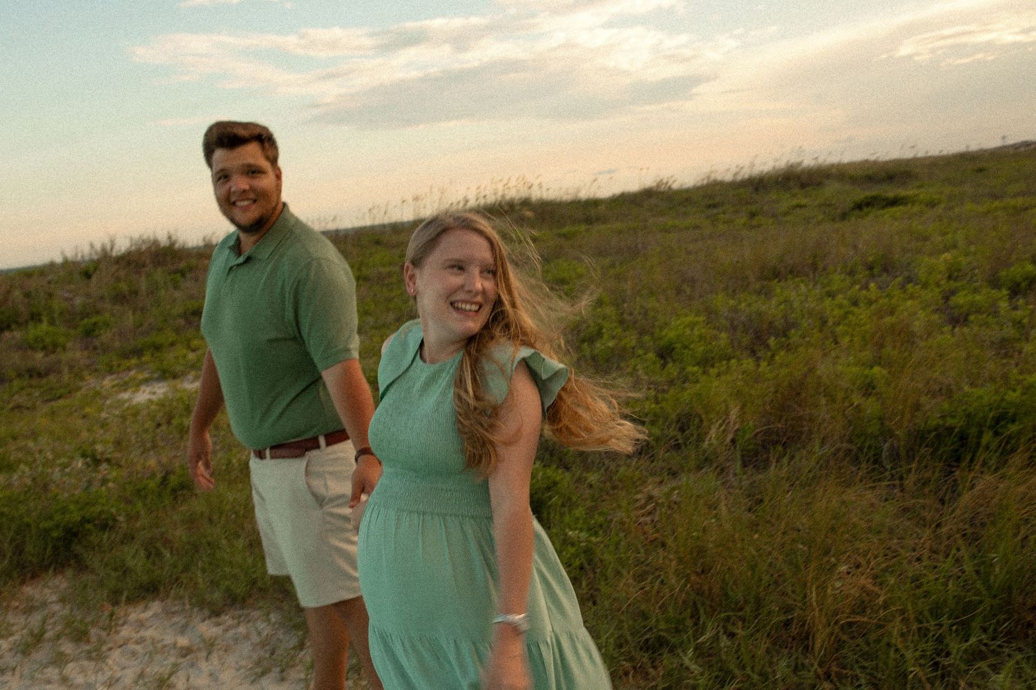 A couple in coordinating mint and khaki outfits share a moment in coastal grasslands.