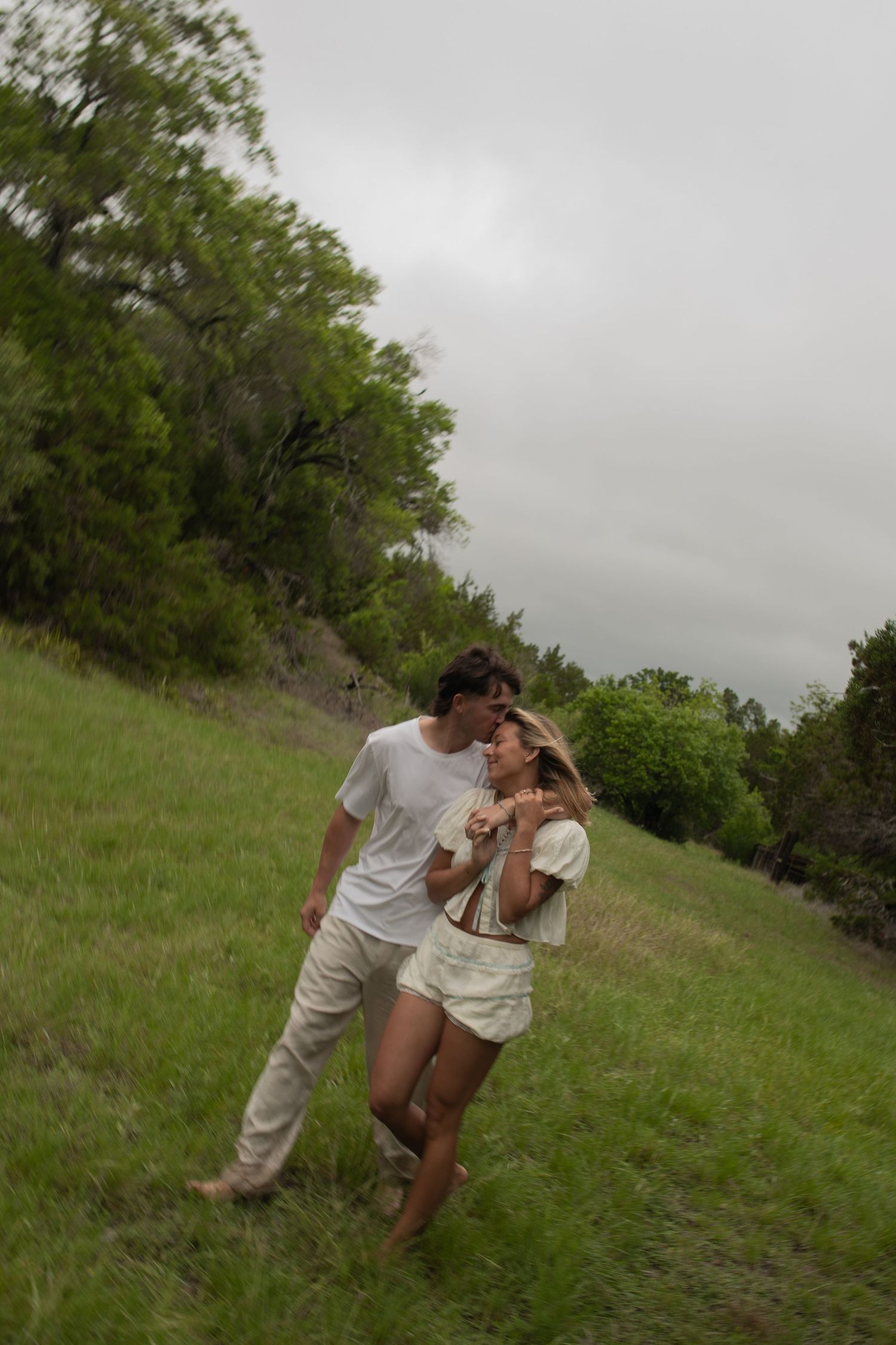 Couple sharing a romantic moment in a lush green meadow, dressed in white, with trees and overcast sky behind them.