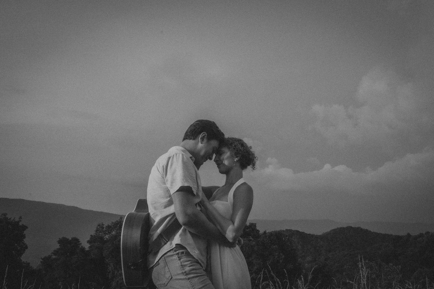 Black and white photo of a romantic couple embracing outdoors with mountains and cloudy sky in the background.