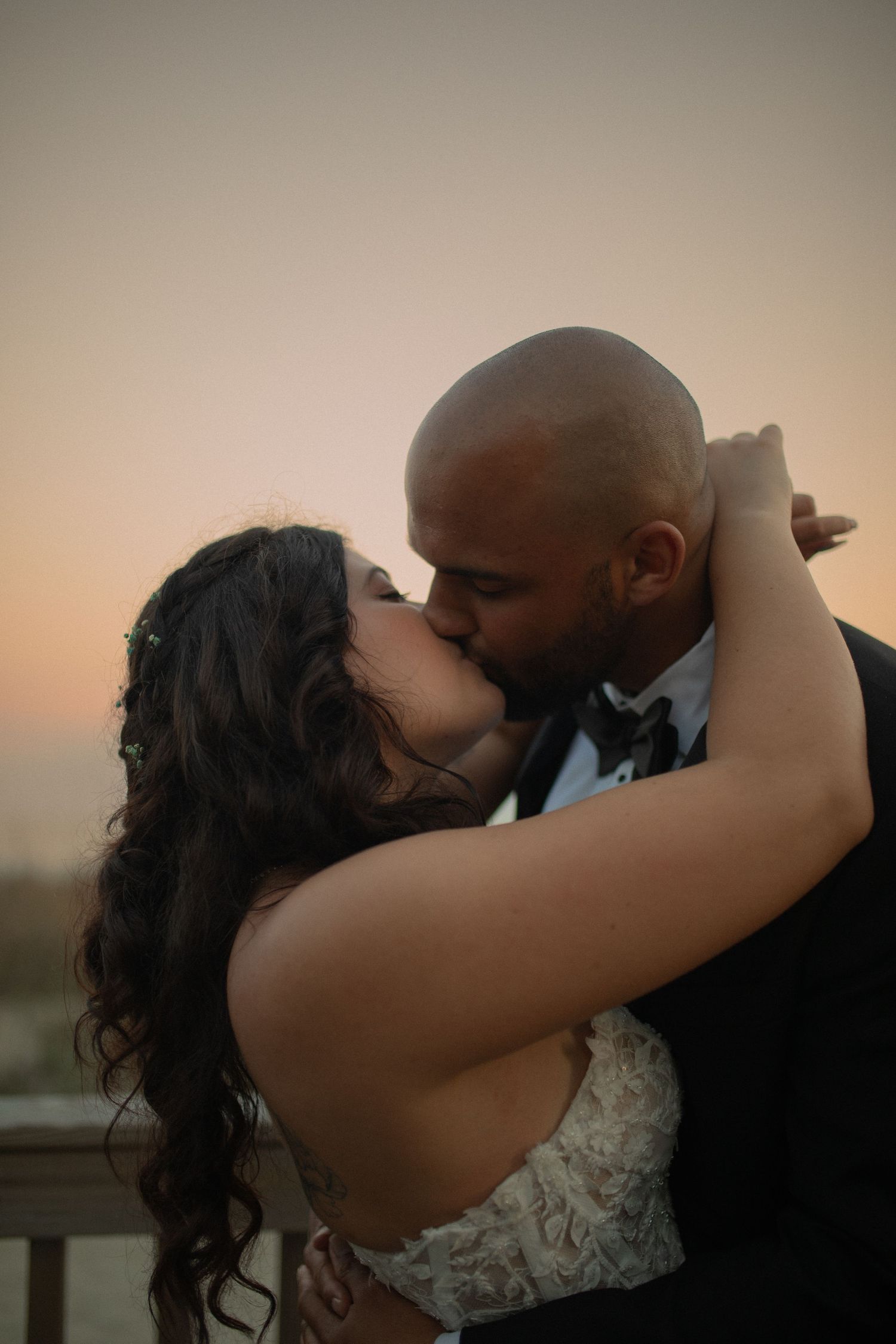Bride and groom share a romantic kiss at sunset, with the bride in a lace gown and groom in a black tuxedo.