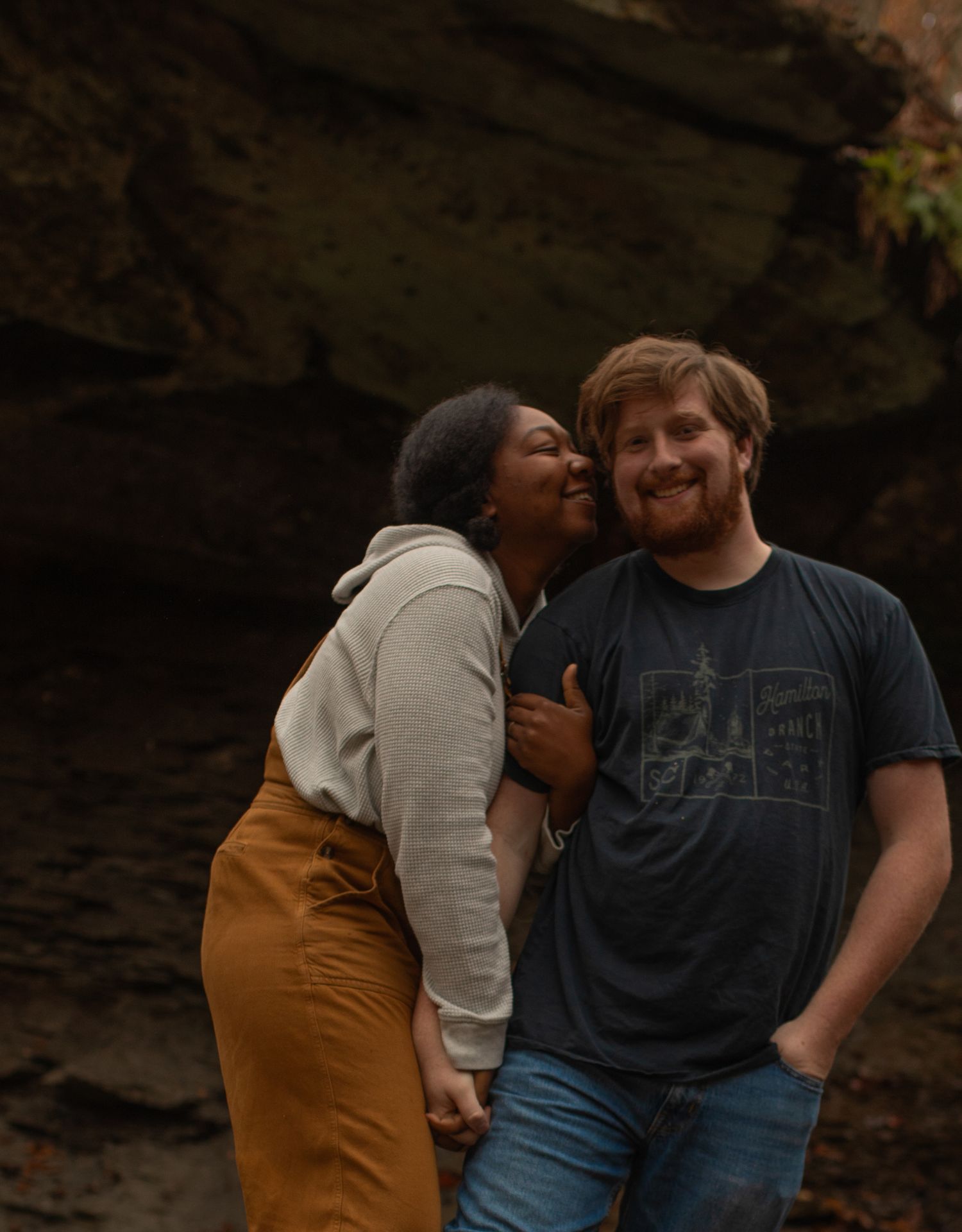 A joyful couple embraces outdoors near rocky cliffs, woman laughing and leaning on smiling bearded man.