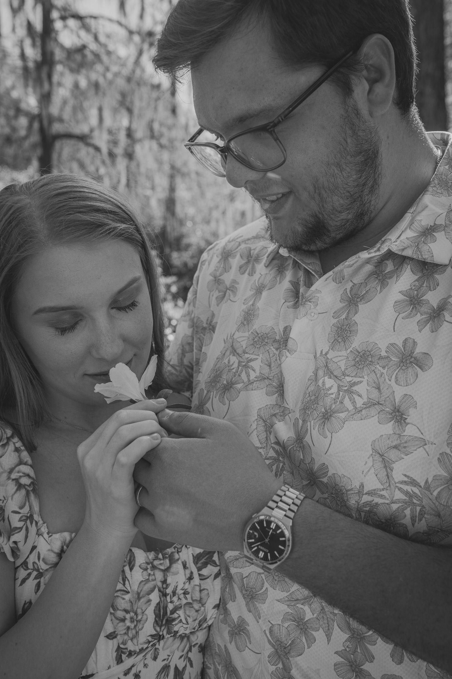 Black and white photo of a couple smelling a flower together outdoors, man wearing glasses and floral shirt.