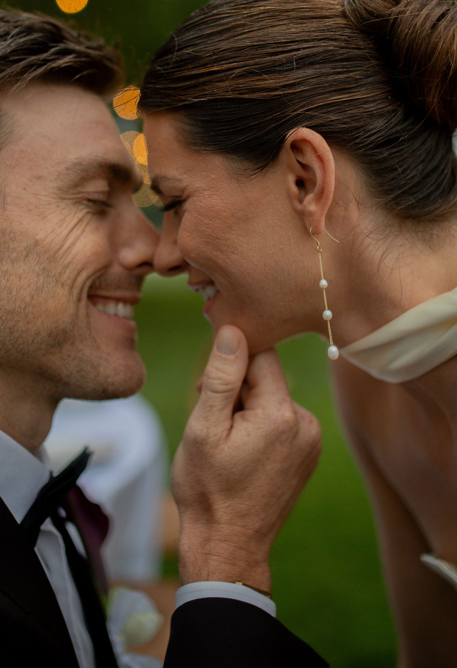 A smiling groom in a tuxedo and bride in a white dress share an intimate forehead-to-forehead moment outdoors.