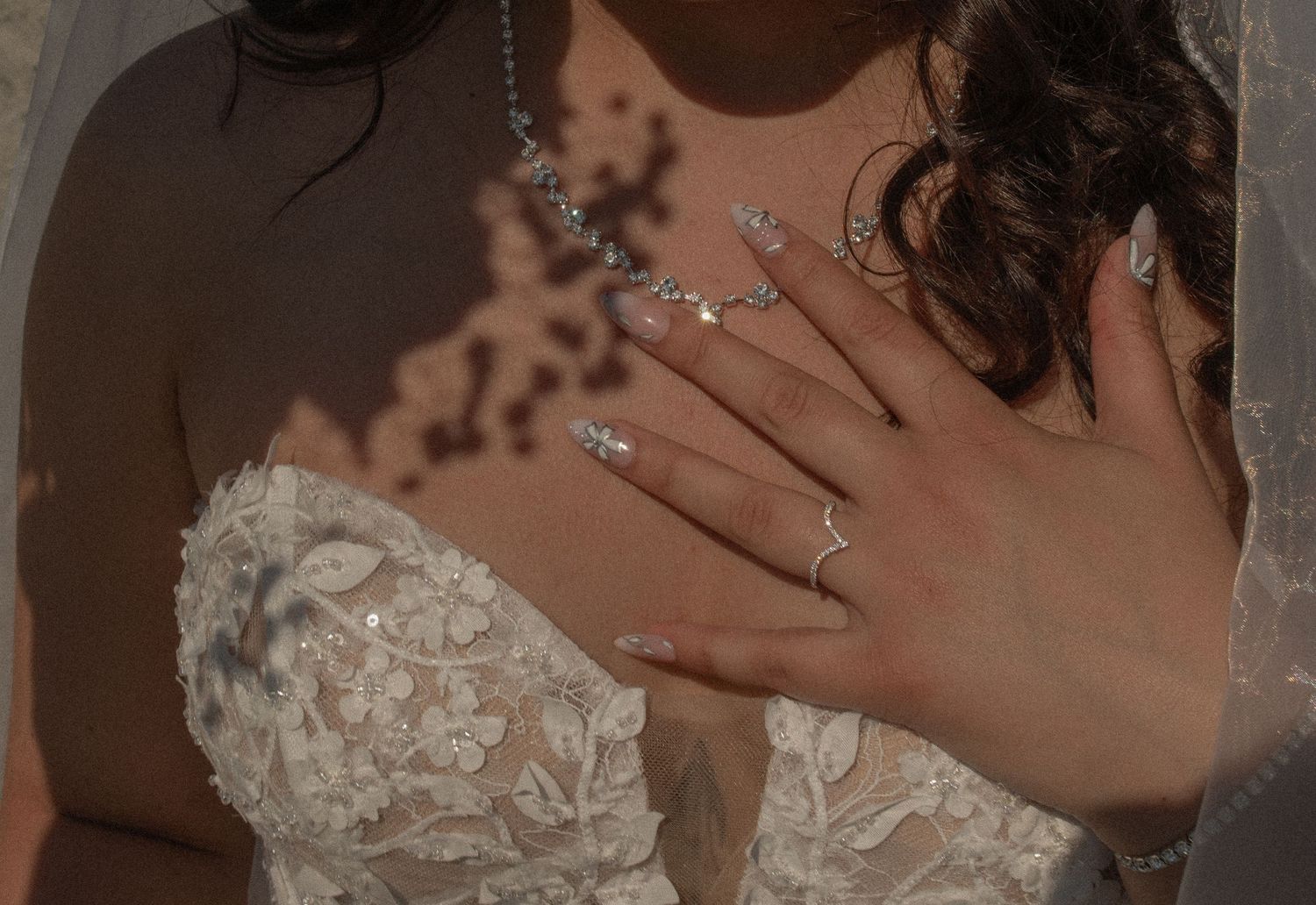 Bride in lace wedding dress with diamond necklace, ring, and elegant rhinestone nail art resting hand on chest.