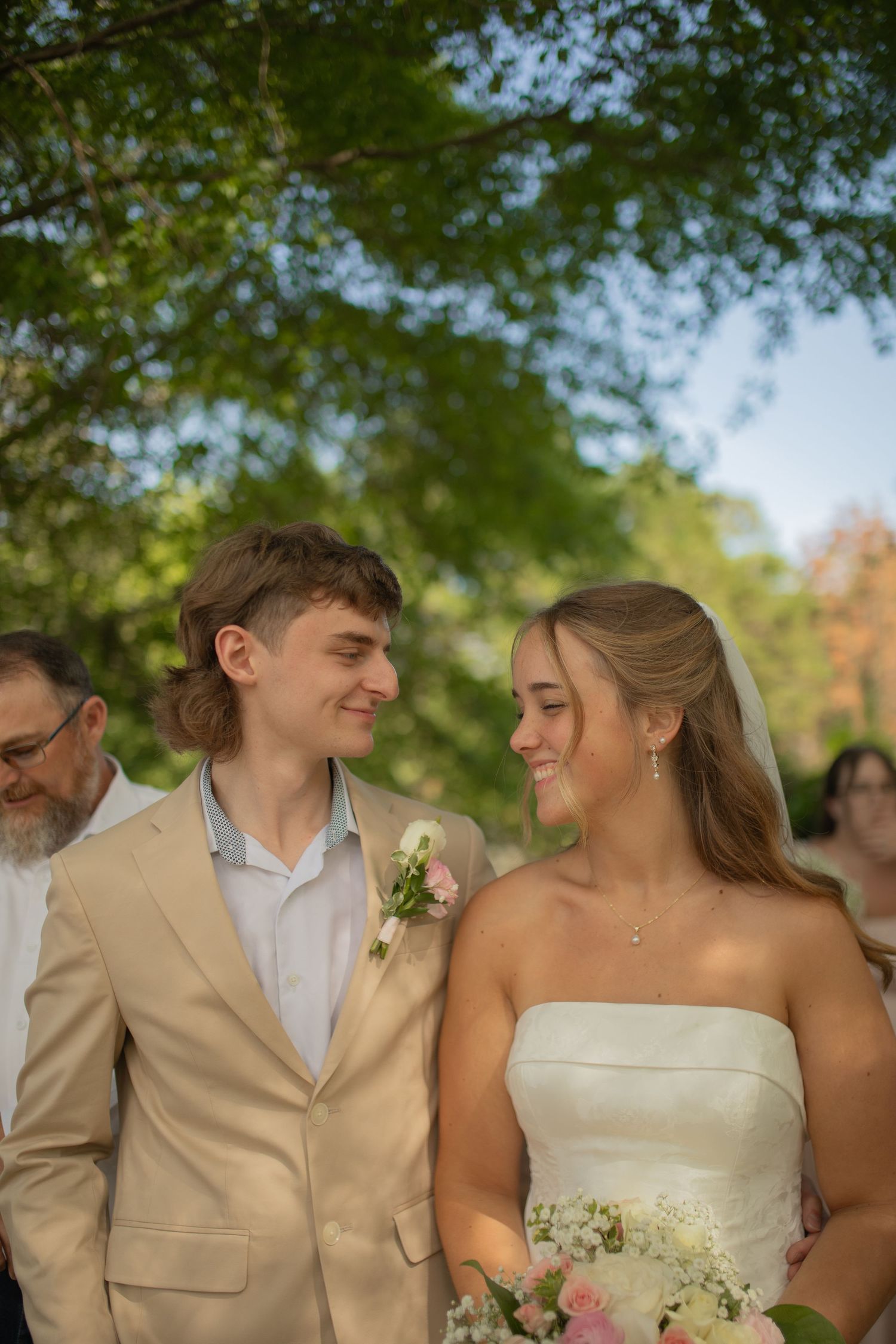 Young couple in wedding attire smiling at each other outdoors under green trees, groom in tan suit, bride in white strapless gown.
