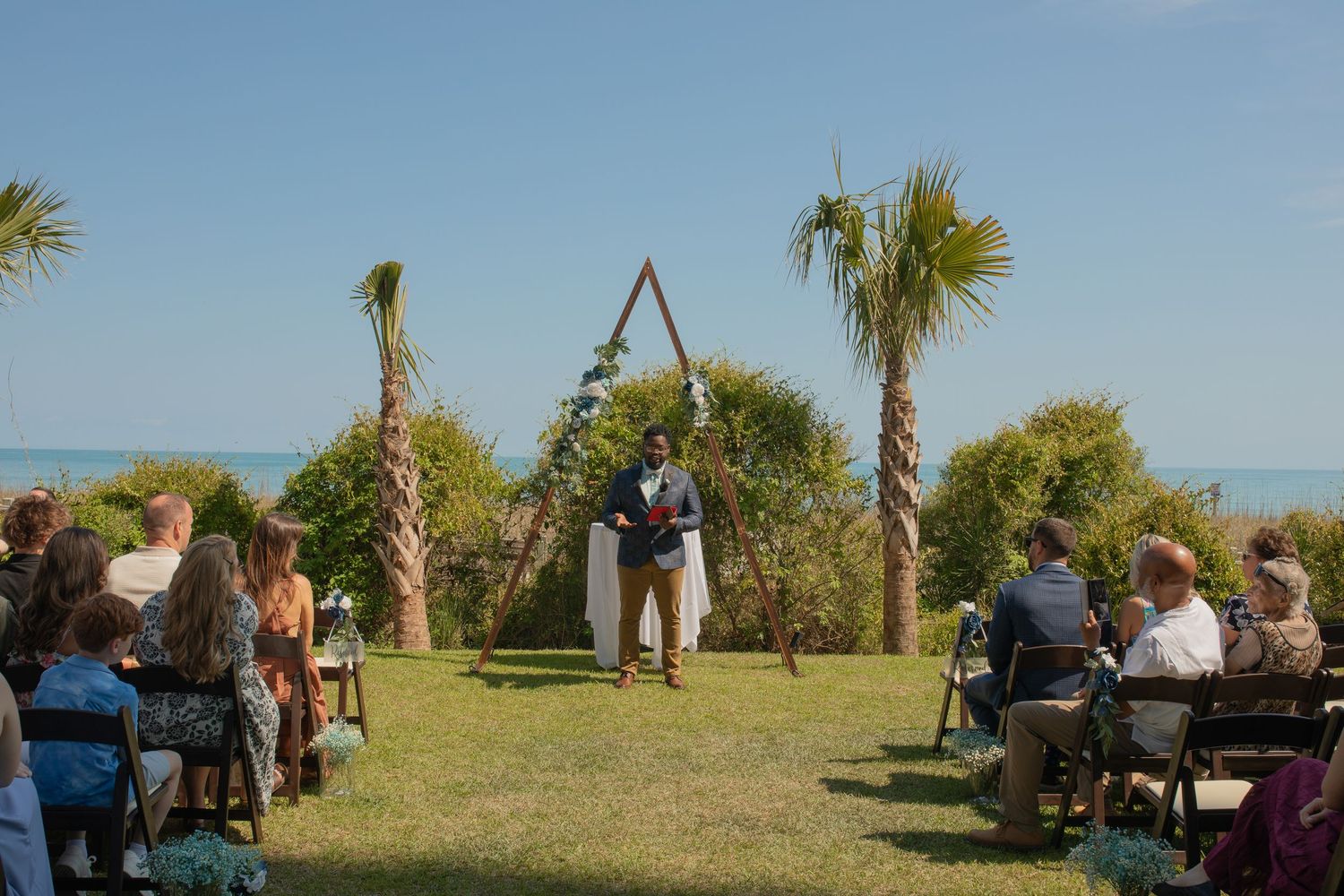 Outdoor beach wedding ceremony with guests seated on lawn, officiant at triangular arch, palm trees and ocean in background.