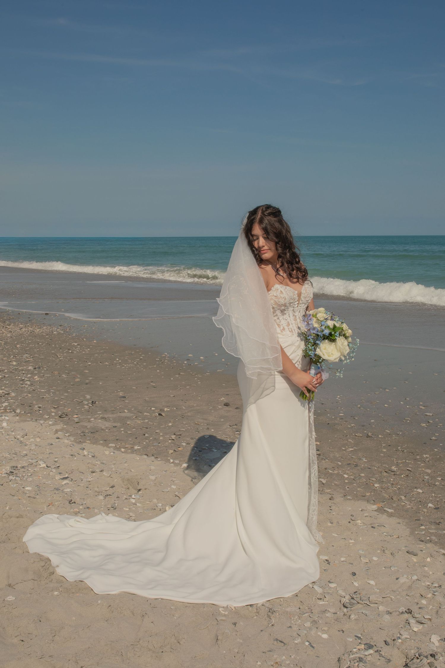 Bride in white gown with veil holds bouquet on sandy beach with ocean waves, perfect for beach wedding inspiration.