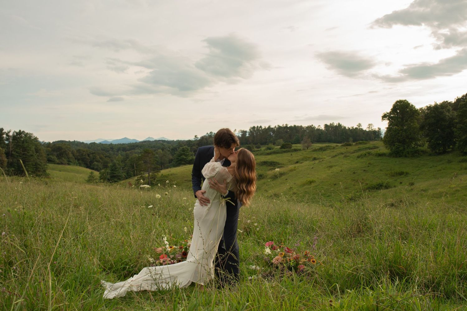 Bride and groom share a romantic kiss in a lush green meadow with rolling hills and cloudy sky backdrop.