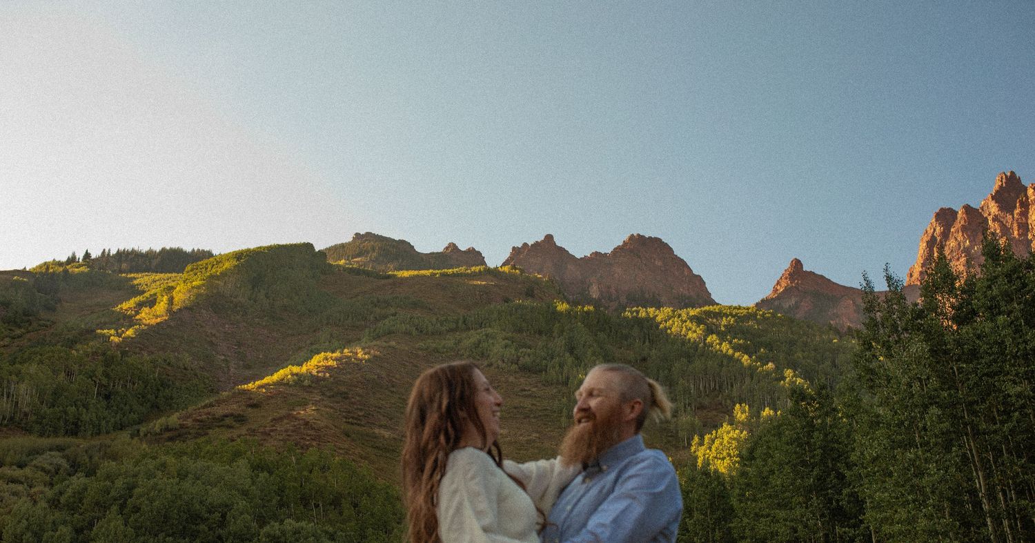 Couple embracing and smiling in front of lush green mountains with golden aspen trees at sunset in Colorado.