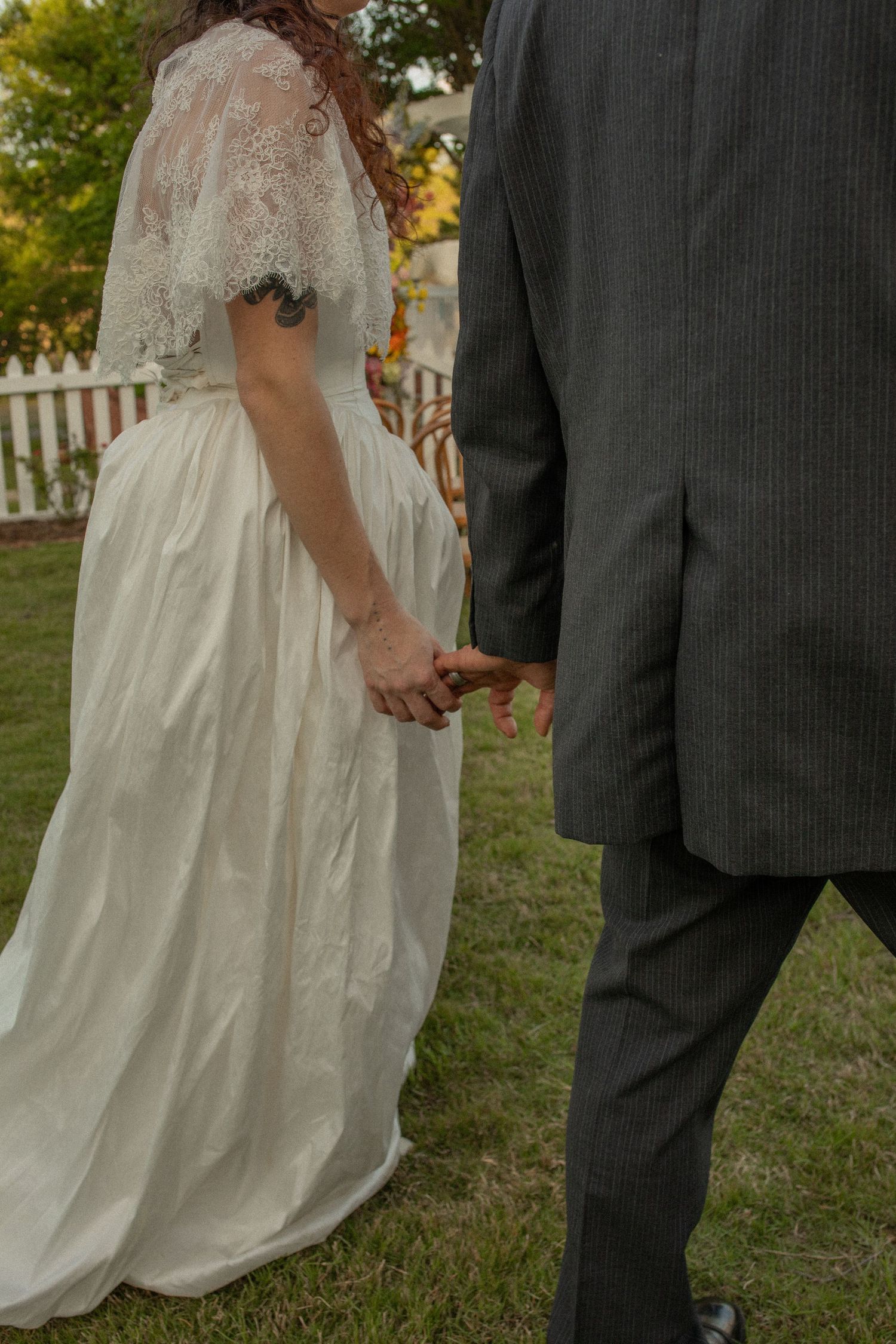 Bride in white lace wedding gown holding hands with groom in gray suit outdoors on green grass.