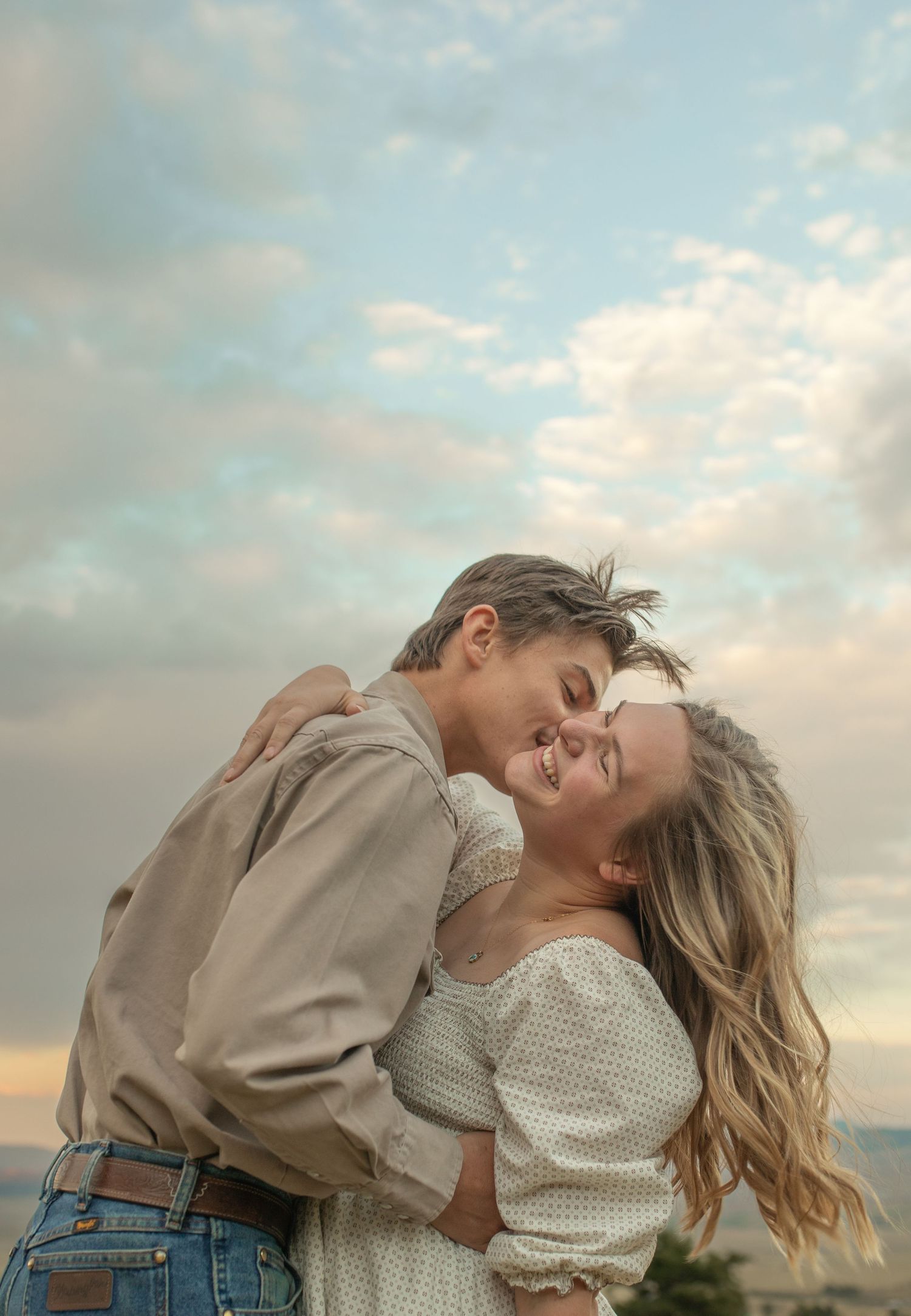 Romantic couple embracing and smiling under a cloudy sky at sunset, sharing a tender moment outdoors.