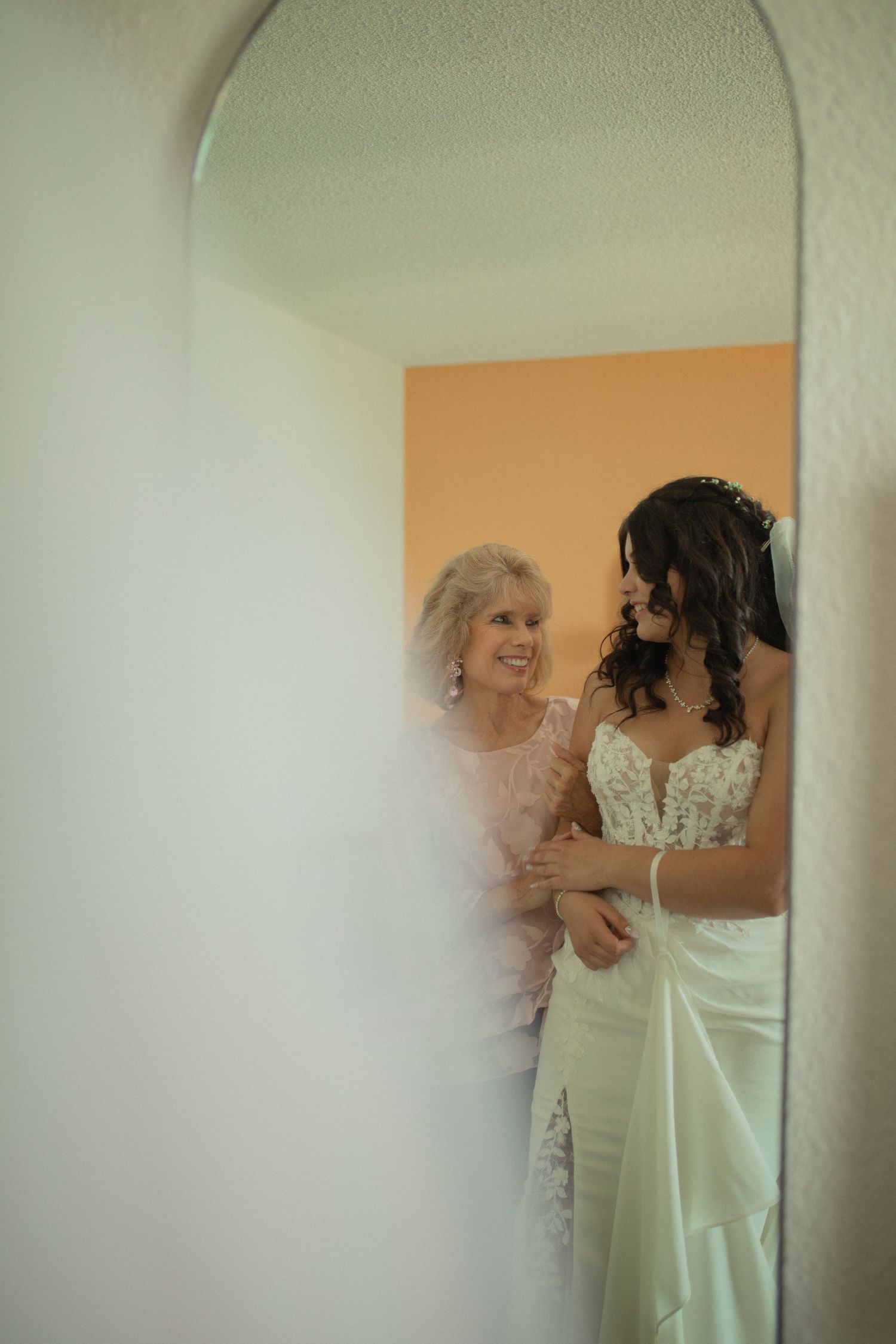 A bride in a white lace gown shares a tender moment with an older woman in a mirror reflection.