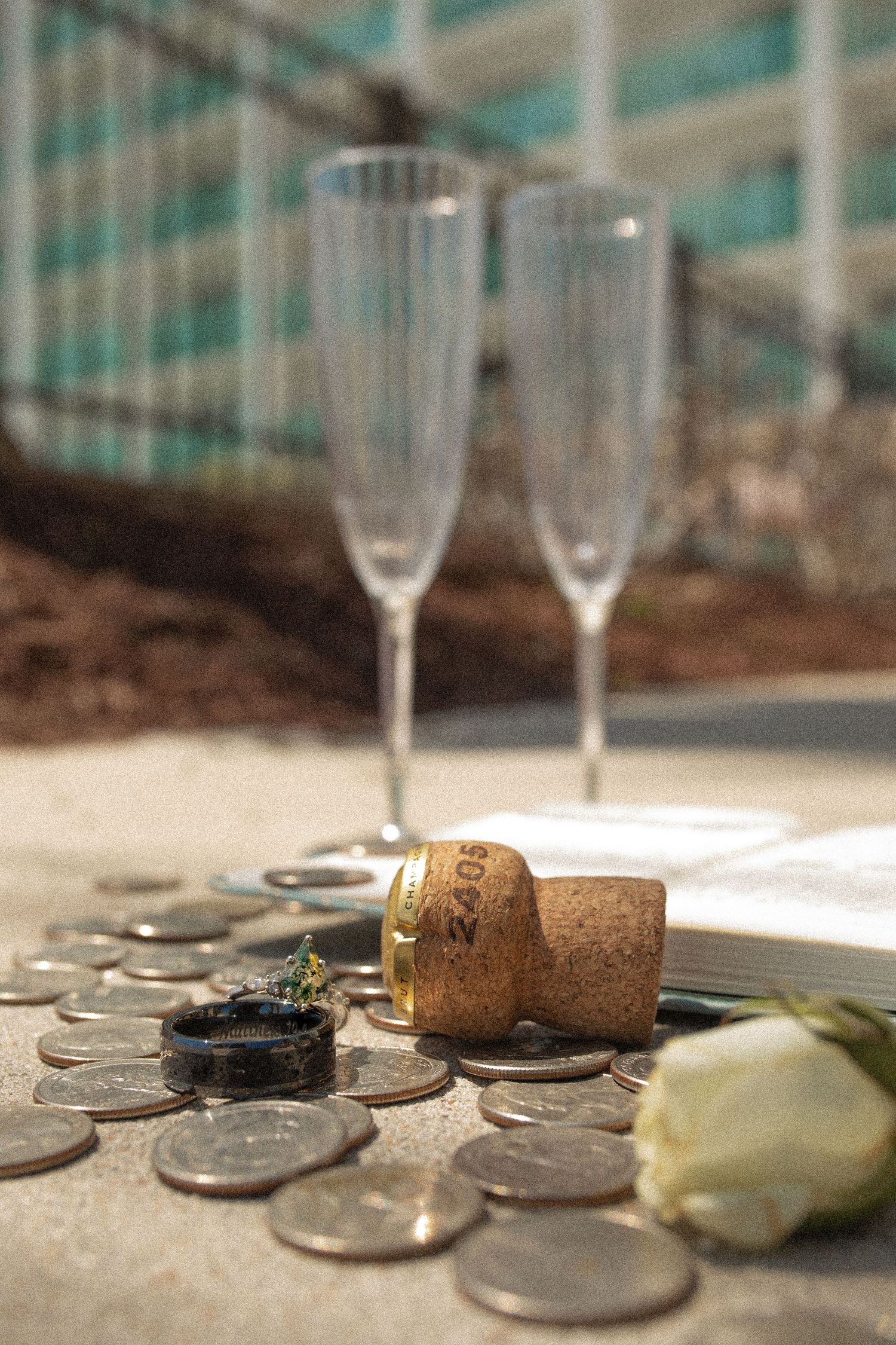 Champagne cork, wedding ring, coins, white rose, and two empty flutes on concrete surface for a celebration.