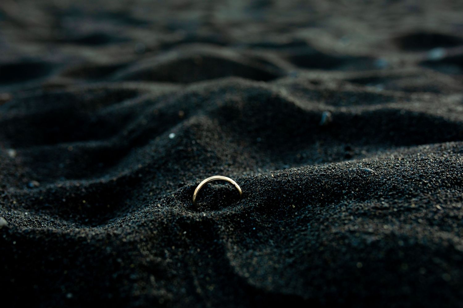 A close-up of a tungsten wedding ring resting on dark textured sand at the beach during sunset.