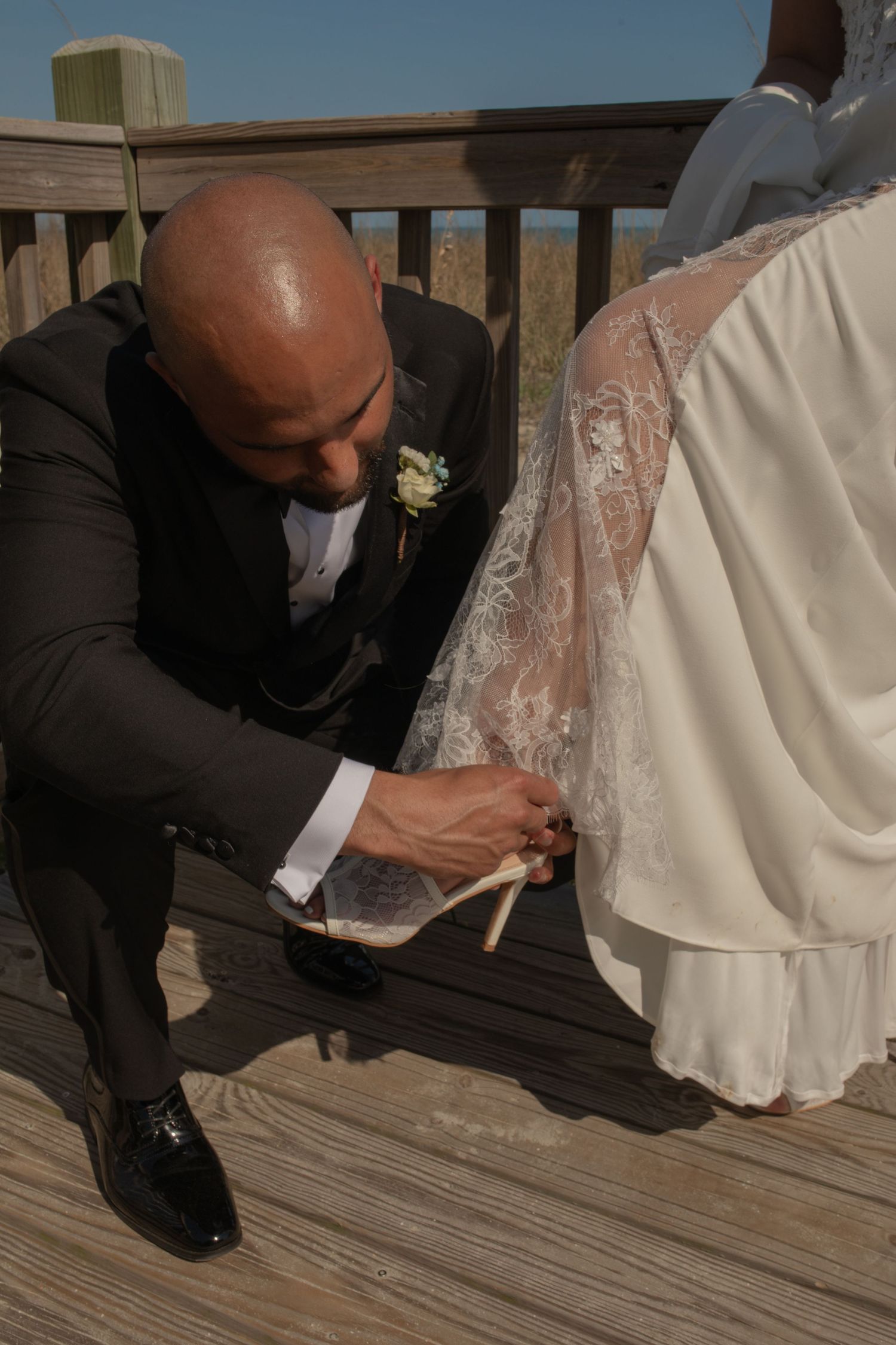 Groom in black suit kneeling on wooden deck to help bride with her wedding shoe during outdoor ceremony.