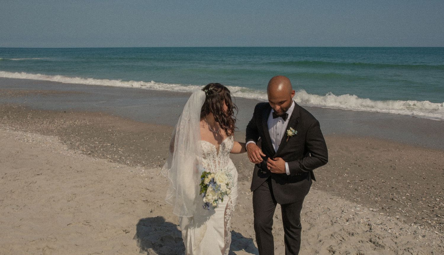 Bride and groom stand on a sandy beach near ocean waves, bride holding white bouquet in wedding dress with veil.