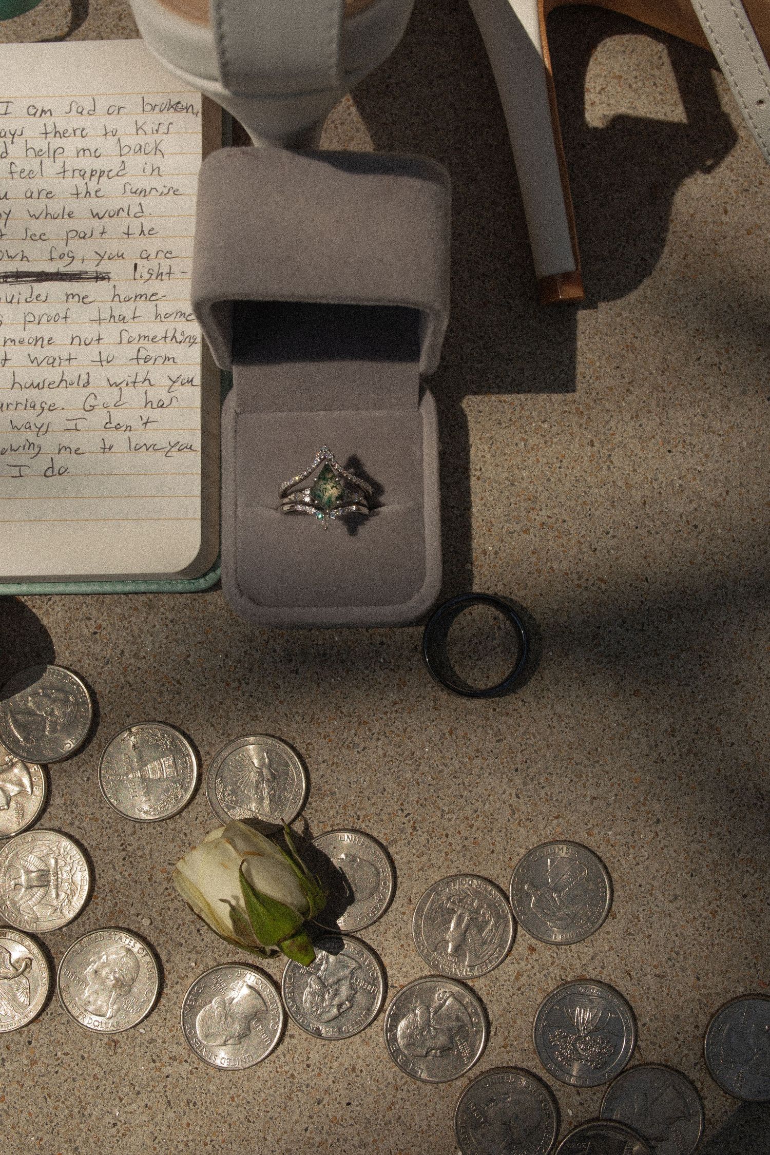 A silver ring with green gemstone in gray velvet box surrounded by coins and a white rose on concrete with handwritten letter.