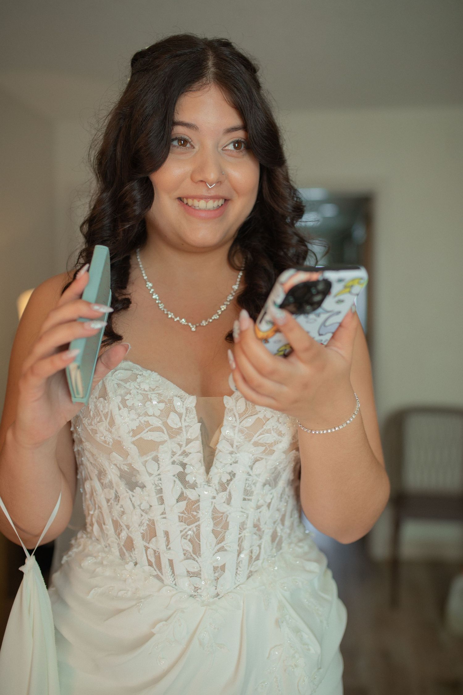 Smiling bride in white lace gown holding two phones, wearing diamond necklace and bracelet on her wedding day.