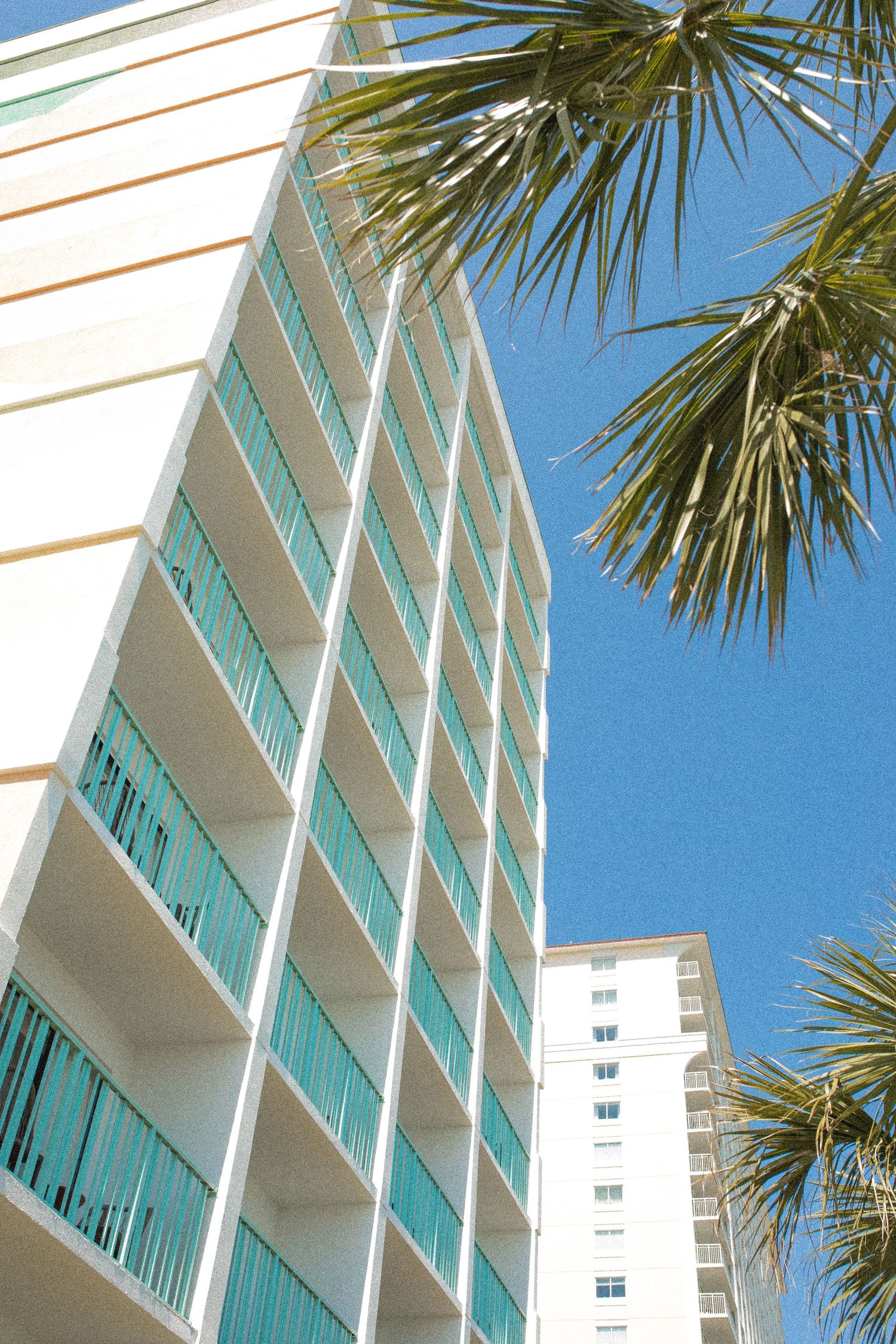 Upward view of a modern beachfront high-rise with turquoise balconies, white facade, and palm trees against a clear blue sky.