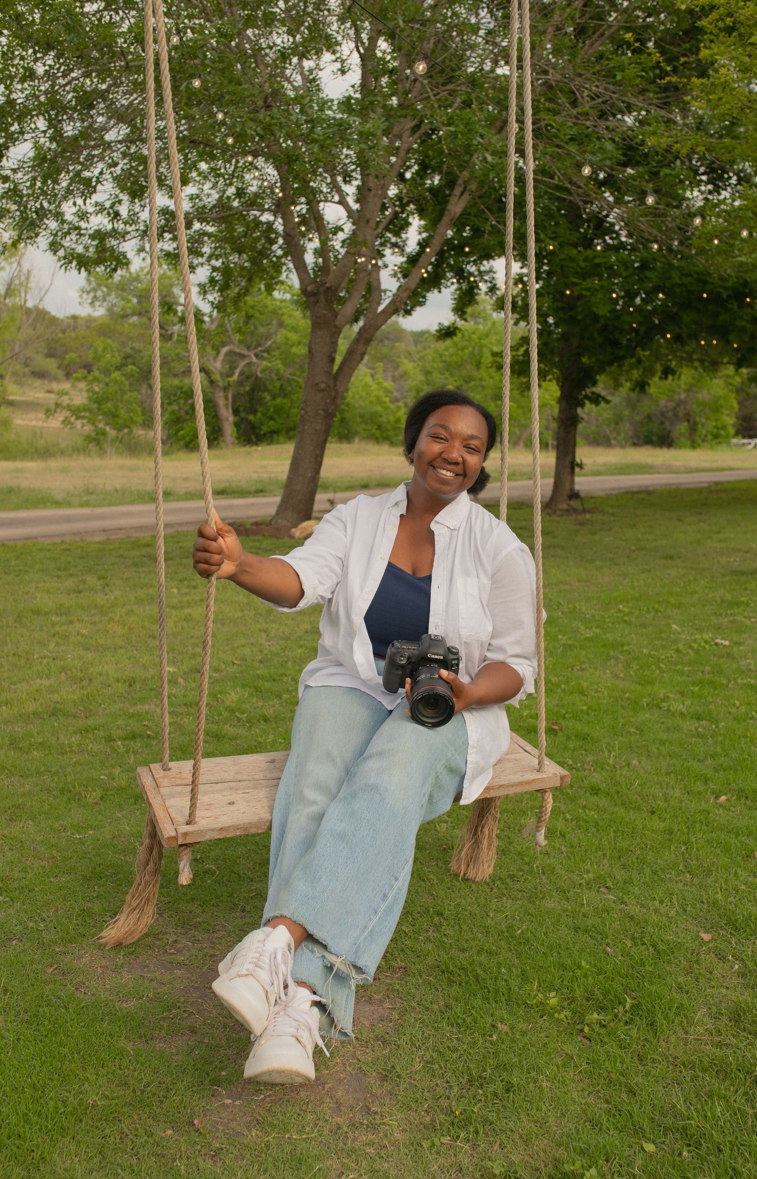 A smiling woman sits on a wooden rope swing outdoors, holding a camera, wearing jeans and a white shirt.