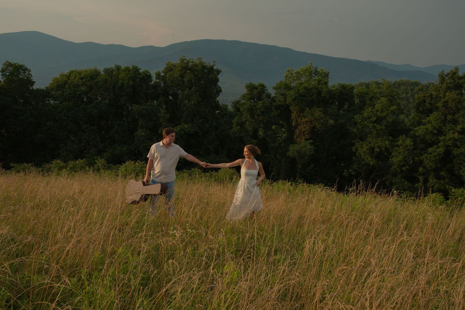 Couple holding hands running through golden meadow with mountain forest backdrop at sunset.