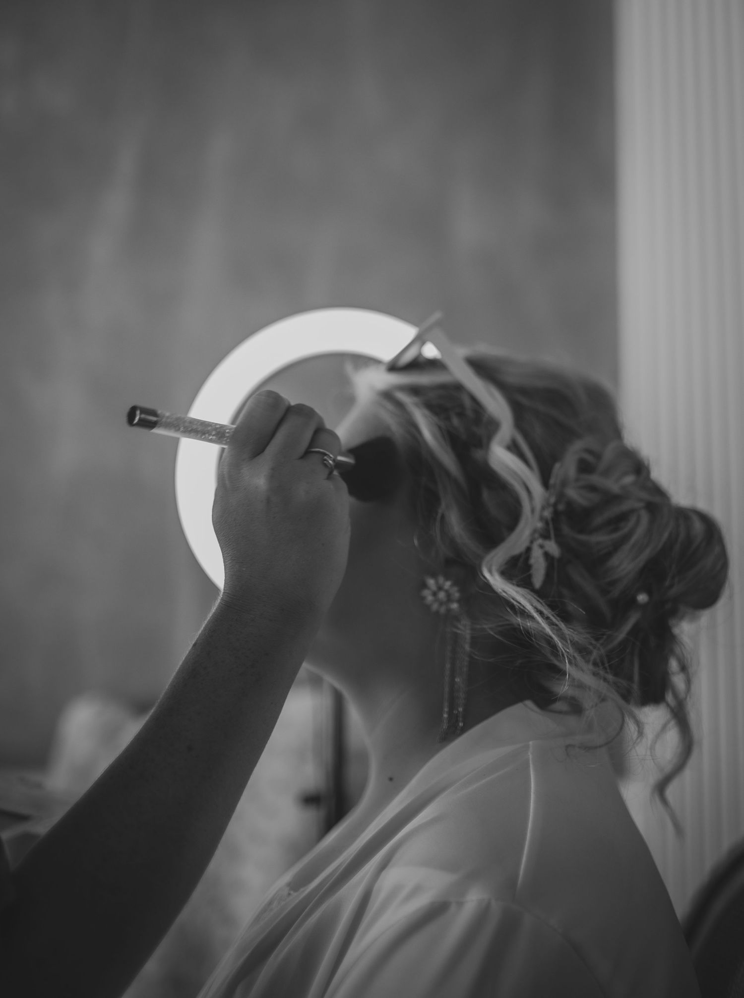 Black and white image showing makeup being applied with a brush while silhouetted against a bright circular light.