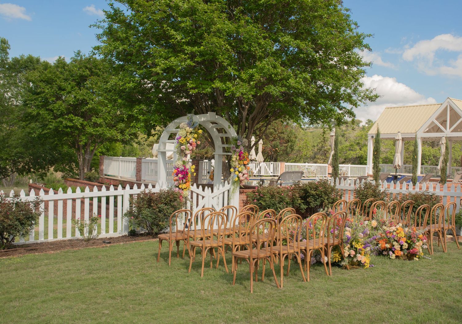 Outdoor wedding ceremony setup with wooden chairs, colorful floral arrangements, white picket fence, and a floral arch on a sunny day.