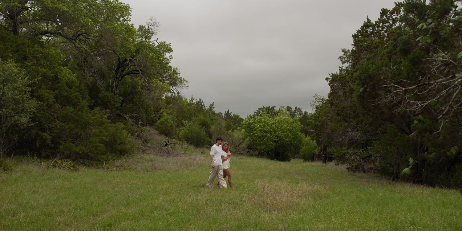 Person walking through a lush green meadow surrounded by dense trees under an overcast sky in a rural landscape.