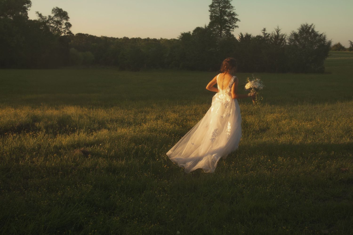 Bride in white wedding gown holding bouquet walks through lush green field at golden hour sunset.