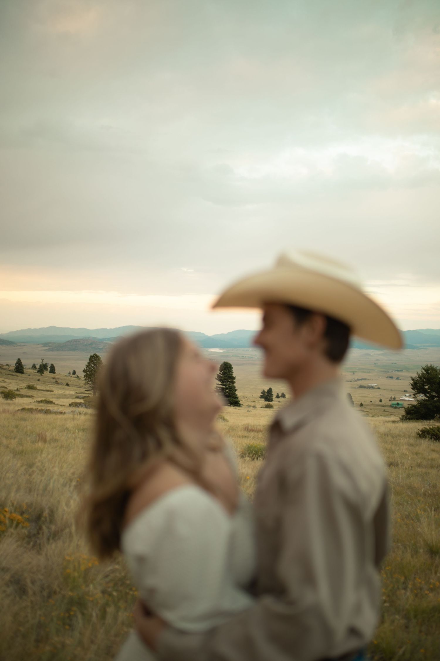 Couple embracing in golden meadow at sunset, man wearing cowboy hat with mountain landscape in background.