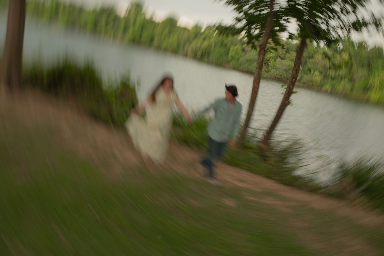 Blurred motion photo of a couple holding hands running along a scenic lakeside path surrounded by lush green trees.