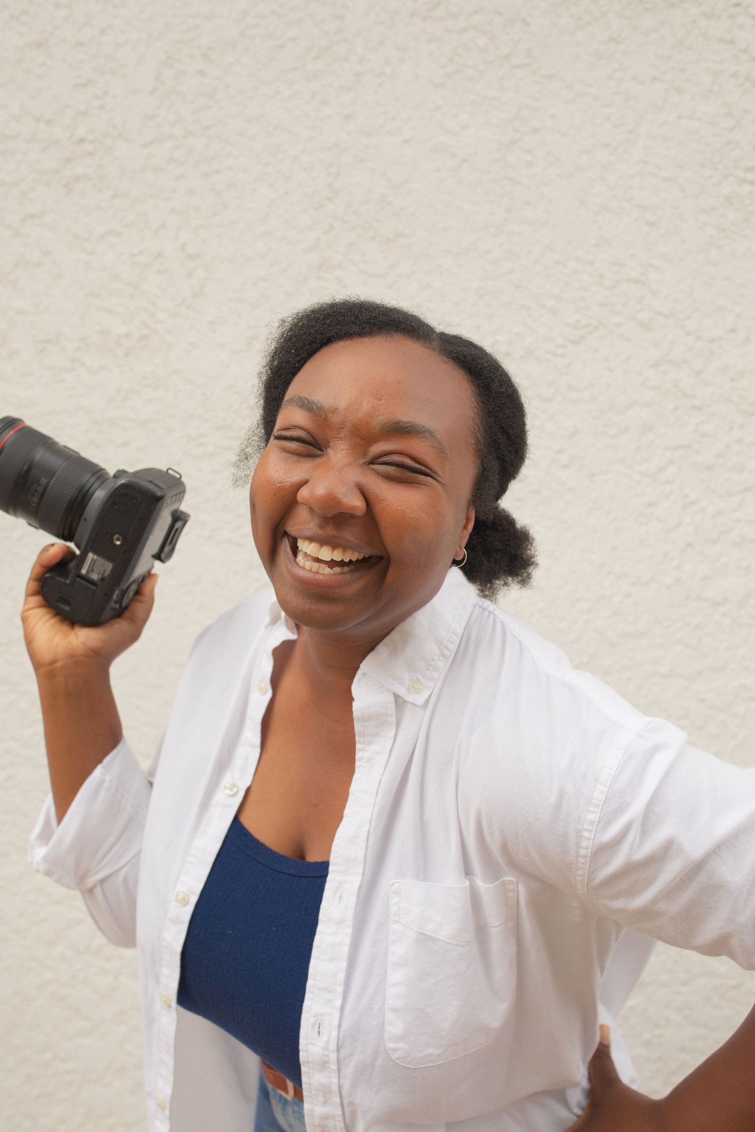 Smiling Black woman holding a DSLR camera, wearing a white shirt over a blue top, standing against a light textured wall.